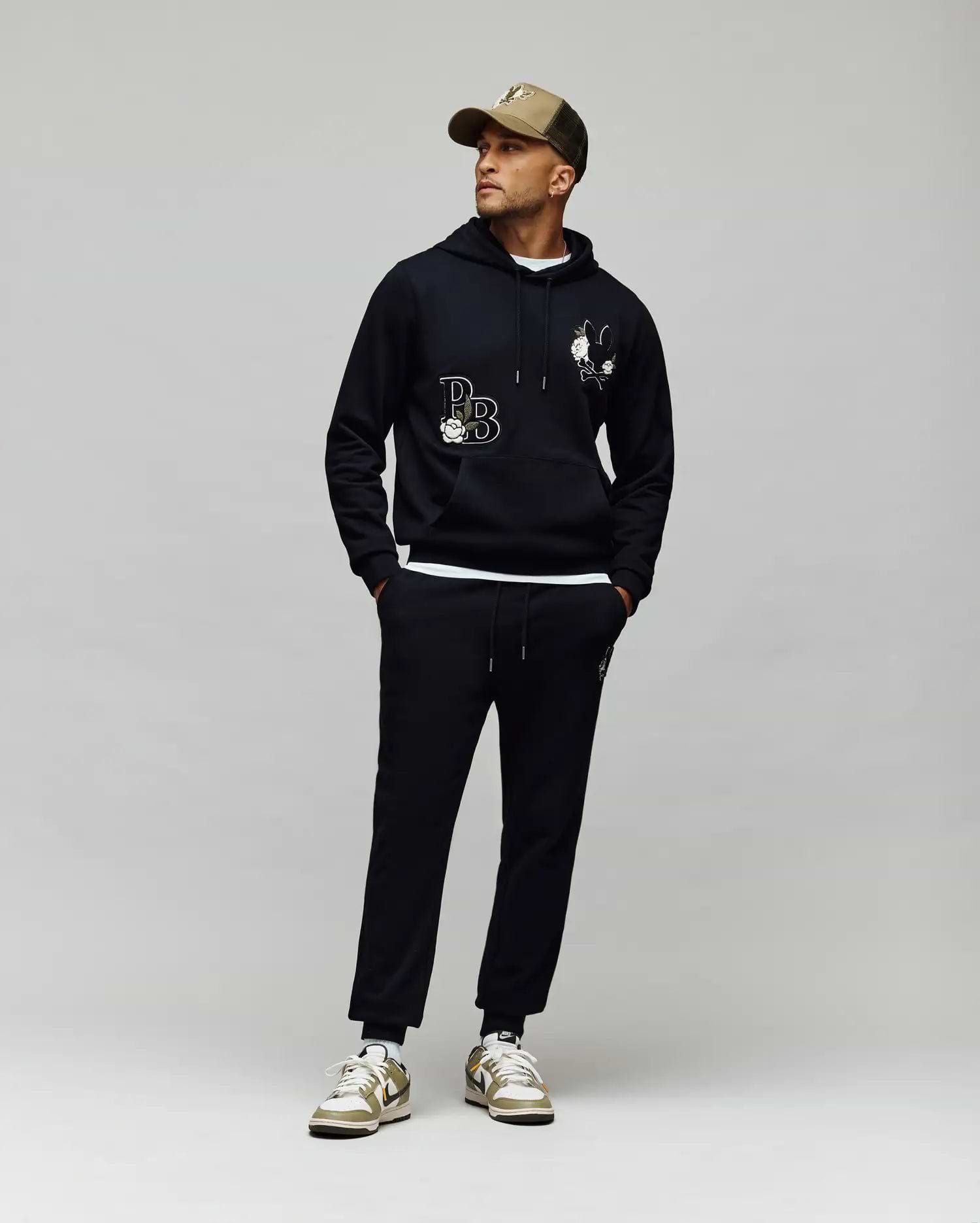 A man in a tan cap, black hoodie with white chenille patches, Psycho Bunny MENS AUGUSTE JOGGER - MB0800015 (100% cotton), and white sneakers with gold accents stands against a light gray background, looking to his right.