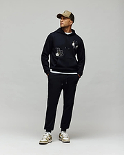 A man in a tan cap, black hoodie with white chenille patches, Psycho Bunny MENS AUGUSTE JOGGER - MB0800015 (100% cotton), and white sneakers with gold accents stands against a light gray background, looking to his right.