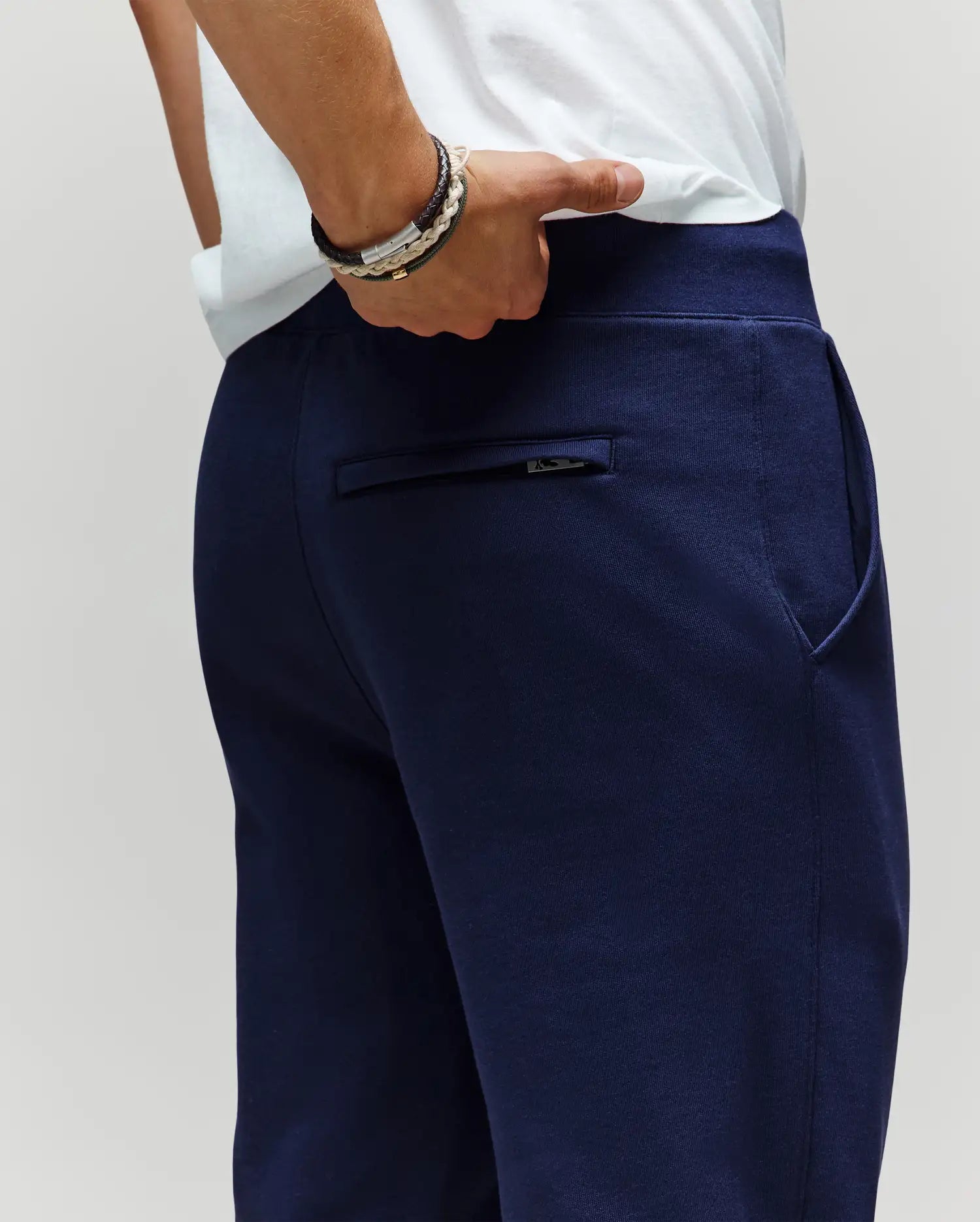 A person in a white shirt and Psycho Bunny MENS AUGUSTE JOGGER - MB0800015 (navy blue, 100% cotton) stands with one hand in their pocket, showcasing the joggers’ back zipper pocket and layered bracelets on one wrist.