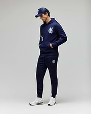 A man stands against a light gray background, hands in his pockets and looking down, wearing Psycho Bunny MENS AUGUSTE JOGGER - MB0800015, a navy cartoon graphic hoodie, navy baseball cap, and white sneakers. Joggers are made from 100% cotton.