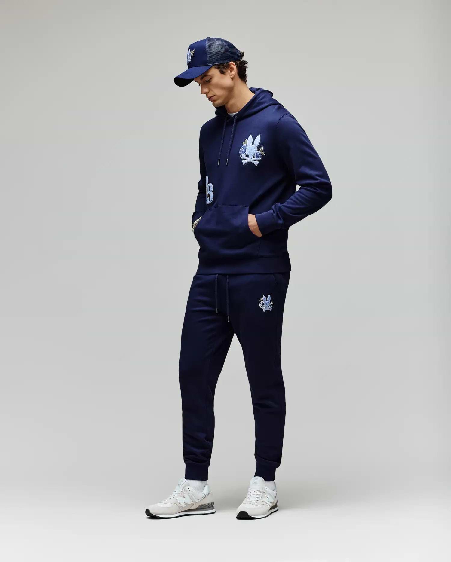 A man stands against a light gray background, hands in his pockets and looking down, wearing Psycho Bunny MENS AUGUSTE JOGGER - MB0800015, a navy cartoon graphic hoodie, navy baseball cap, and white sneakers. Joggers are made from 100% cotton.