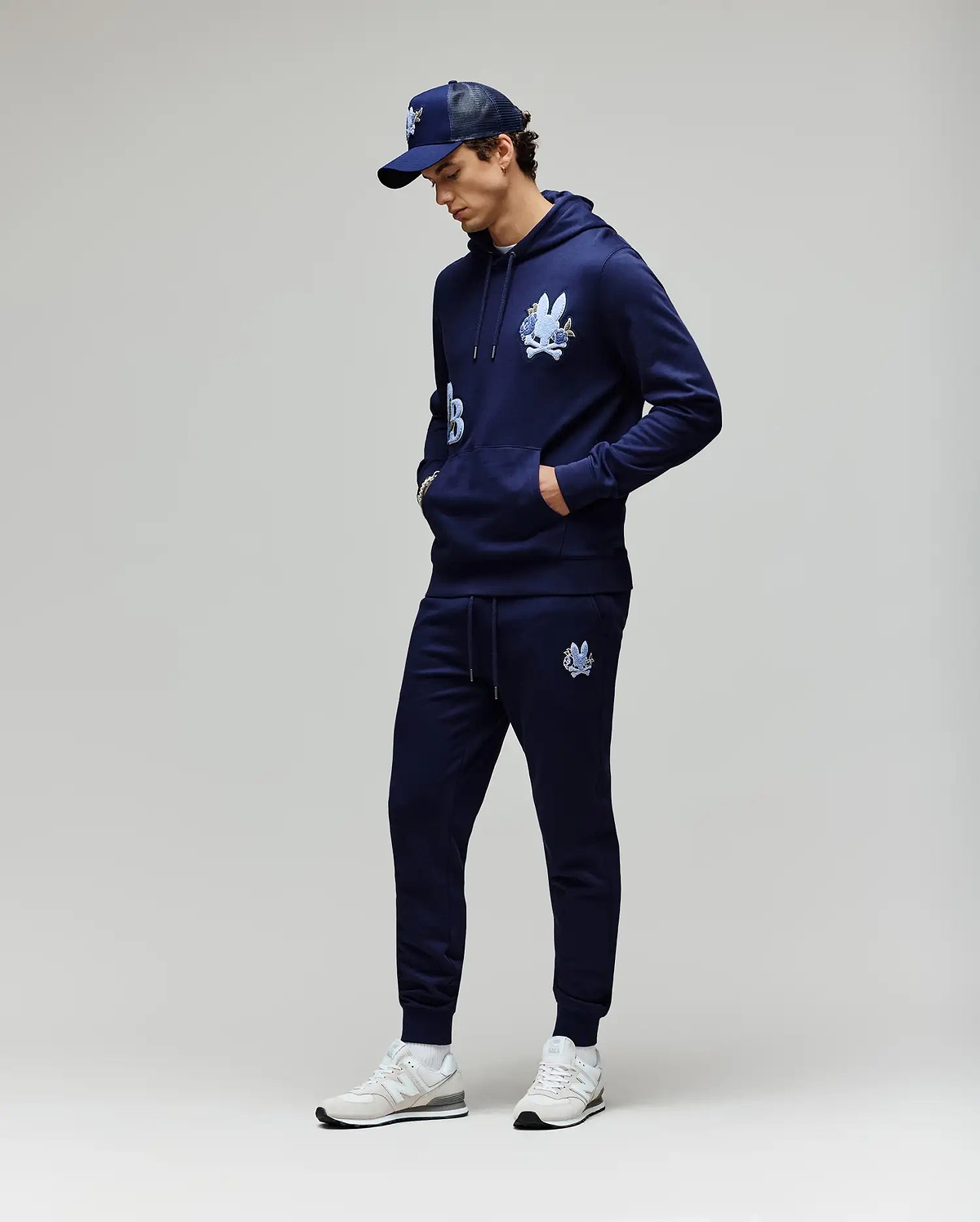 A man stands against a light gray background, hands in his pockets and looking down, wearing Psycho Bunny MENS AUGUSTE JOGGER - MB0800015, a navy cartoon graphic hoodie, navy baseball cap, and white sneakers. Joggers are made from 100% cotton.