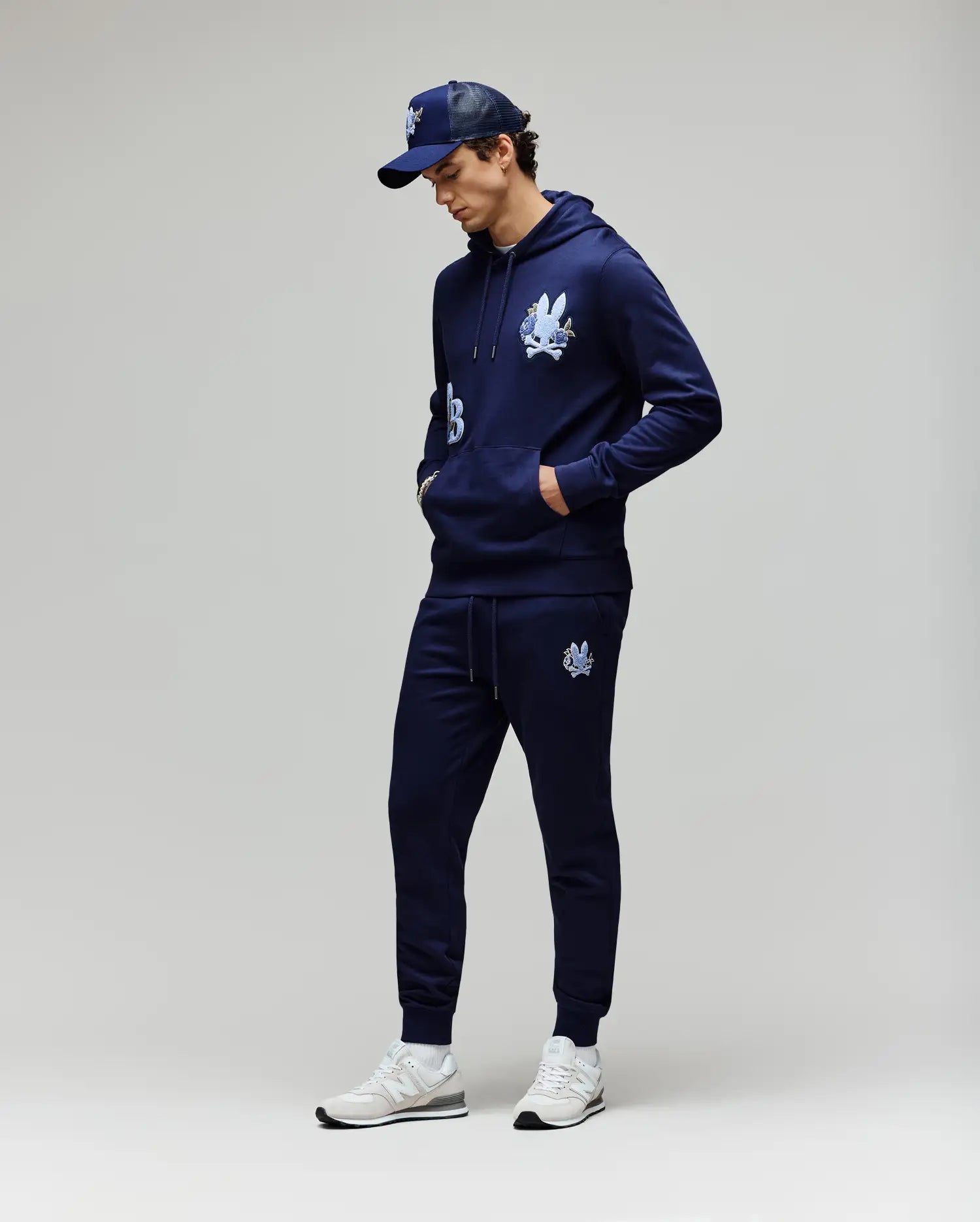 A man stands against a light gray background, hands in his pockets and looking down, wearing Psycho Bunny MENS AUGUSTE JOGGER - MB0800015, a navy cartoon graphic hoodie, navy baseball cap, and white sneakers. Joggers are made from 100% cotton.