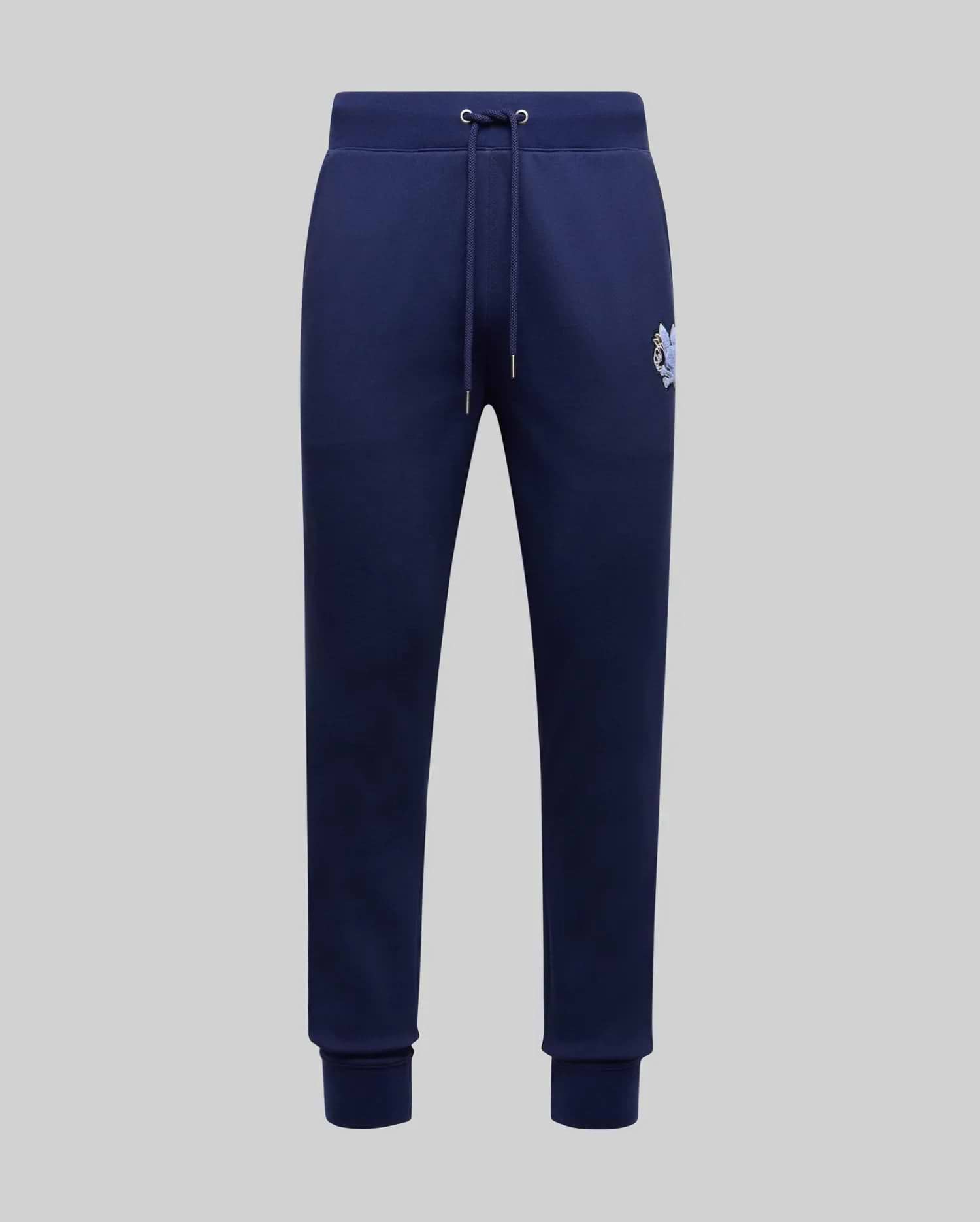 The Psycho Bunny MENS AUGUSTE JOGGER - MB0800015 in navy blue features 100% cotton, a drawstring waistband, side pockets, ribbed cuffs, and a small embroidered logo on the upper left thigh, shown on a light gray background.