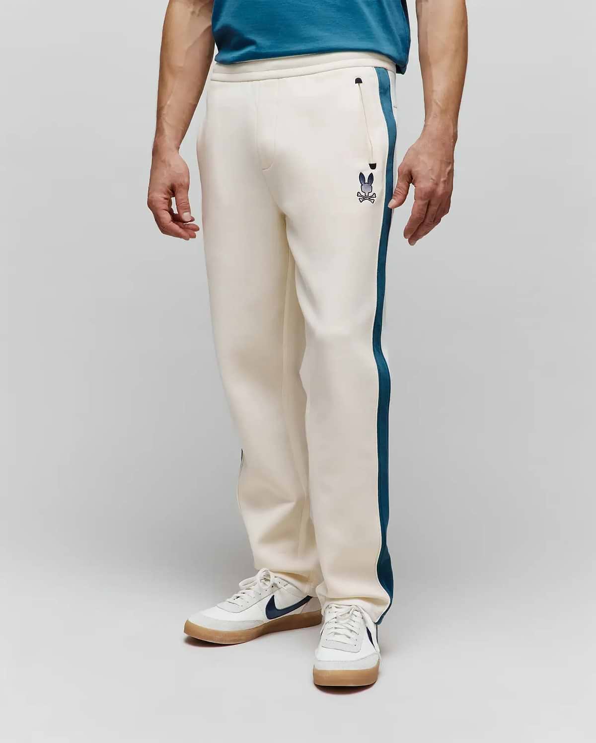 Mens Caspian Track Pant in cream, featuring a relaxed fit, smooth knit fabric, and a dark teal side stripe. A small embroidered Psycho Bunny skull logo is on the left thigh near a zippered pocket. (1)