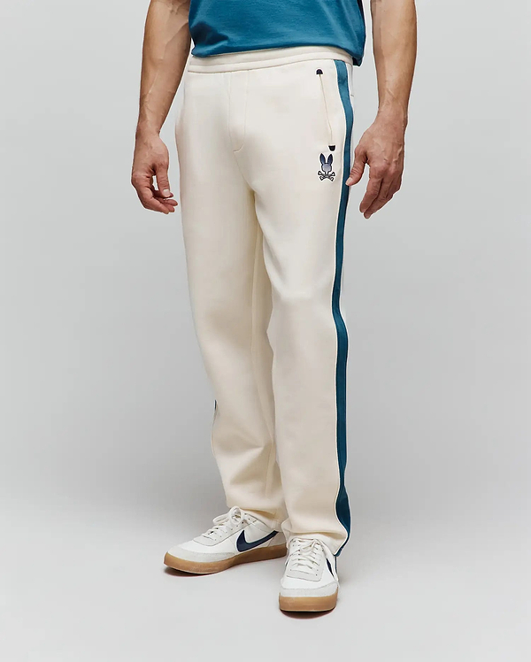 Mens Caspian Track Pant in cream, featuring a relaxed fit, smooth knit fabric, and a dark teal side stripe. A small embroidered Psycho Bunny skull logo is on the left thigh near a zippered pocket.
