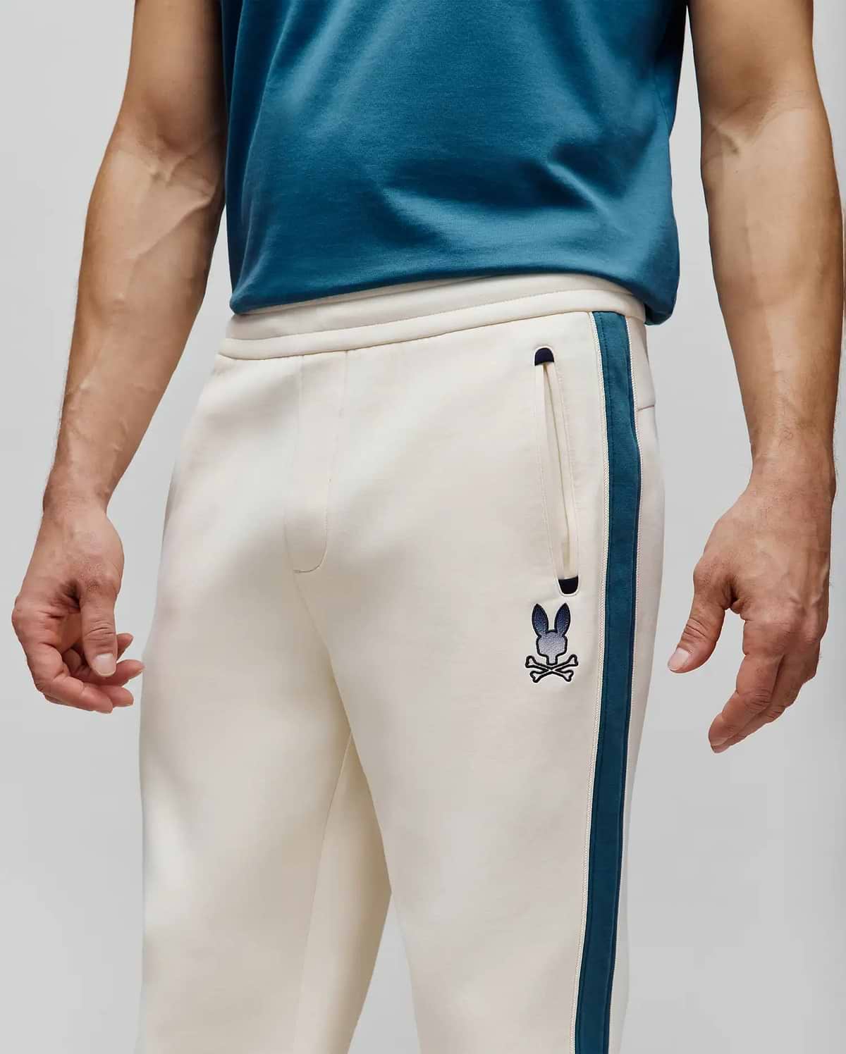 Mens Caspian Track Pant in cream French terry, relaxed fit, with a deep teal side stripe, small embroidered Psycho Bunny skull logo on left thigh, and a zippered pocket. (2)