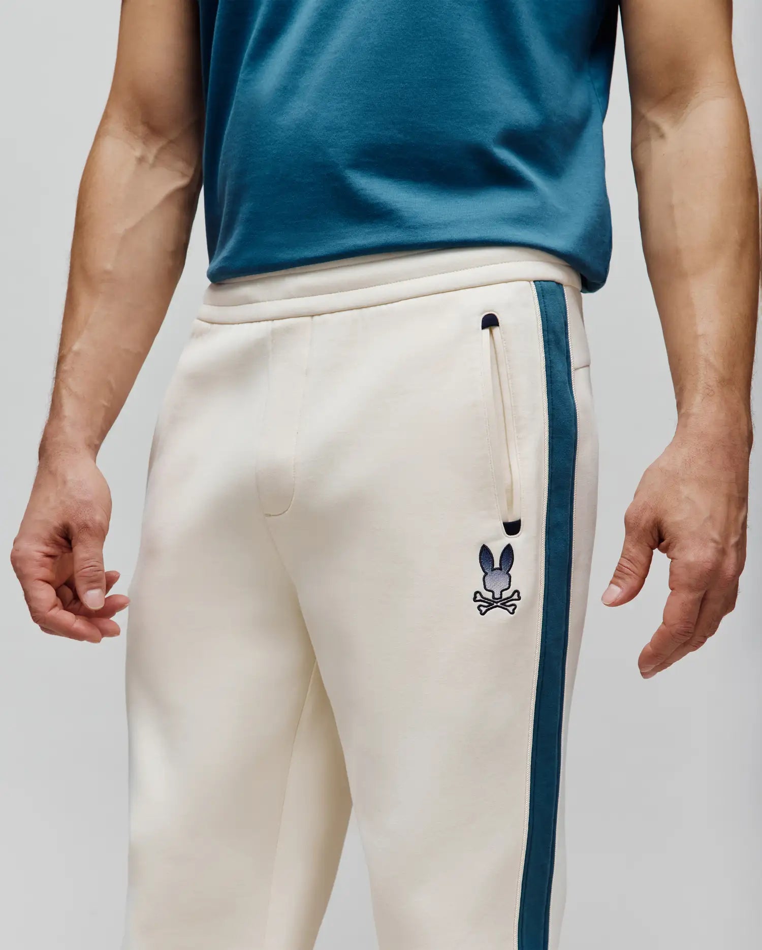 Mens Caspian Track Pant in cream French terry, relaxed fit, with a deep teal side stripe, small embroidered Psycho Bunny skull logo on left thigh, and a zippered pocket. (2)