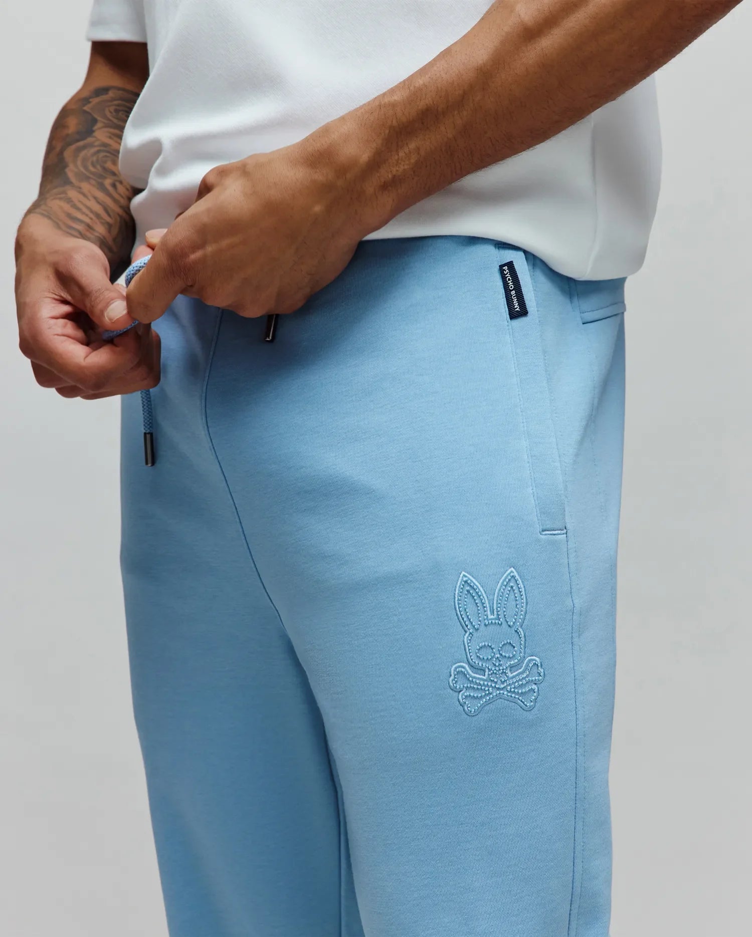 Light blue Mens Barry Joggers MB0800050 in soft jersey knit, featuring a relaxed fit, drawstring waist, and an embroidered tonal Psycho Bunny skull logo on the left thigh, plus a woven brand tag. (2)