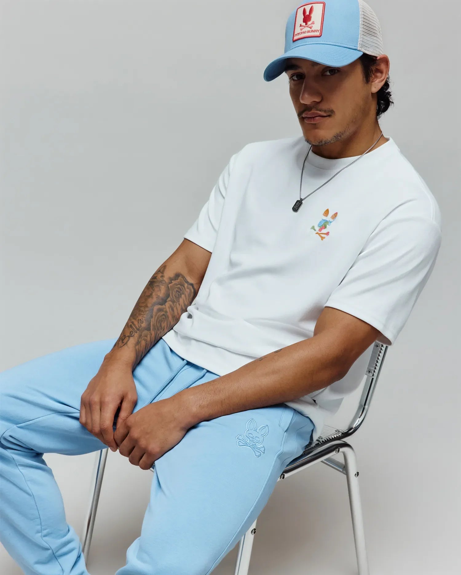 White relaxed fit jersey t-shirt with colorful embroidered Psycho Bunny skull logo, light blue Mens Barry Joggers with tonal bunny skull on left thigh, and blue mesh trucker hat with red logo patch. (4)