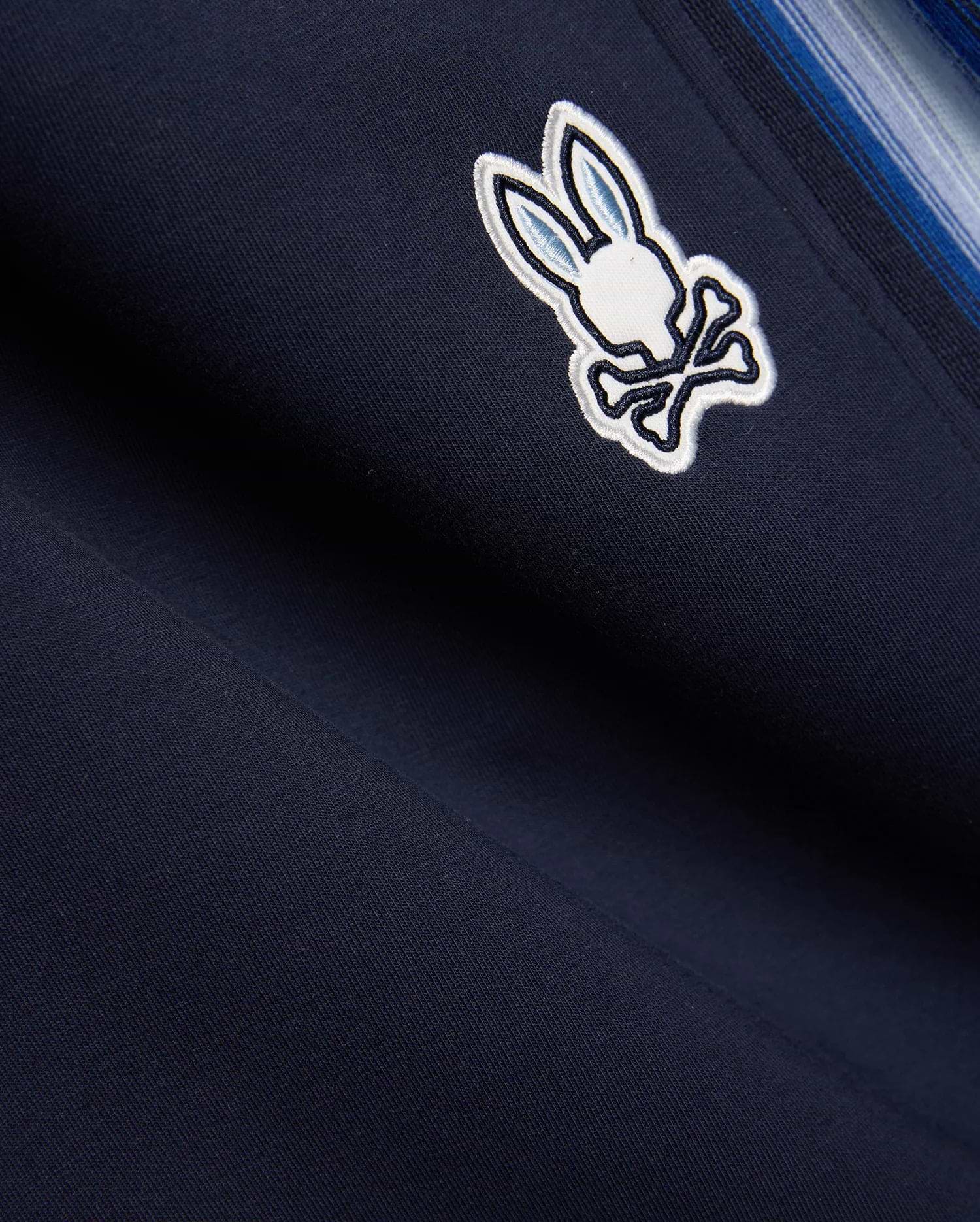 Navy blue Mens Jaydon Track Pant in smooth knit, featuring a prominent embroidered white and light blue Psycho Bunny skull logo and a multi-tonal blue vertical side stripe. (6)