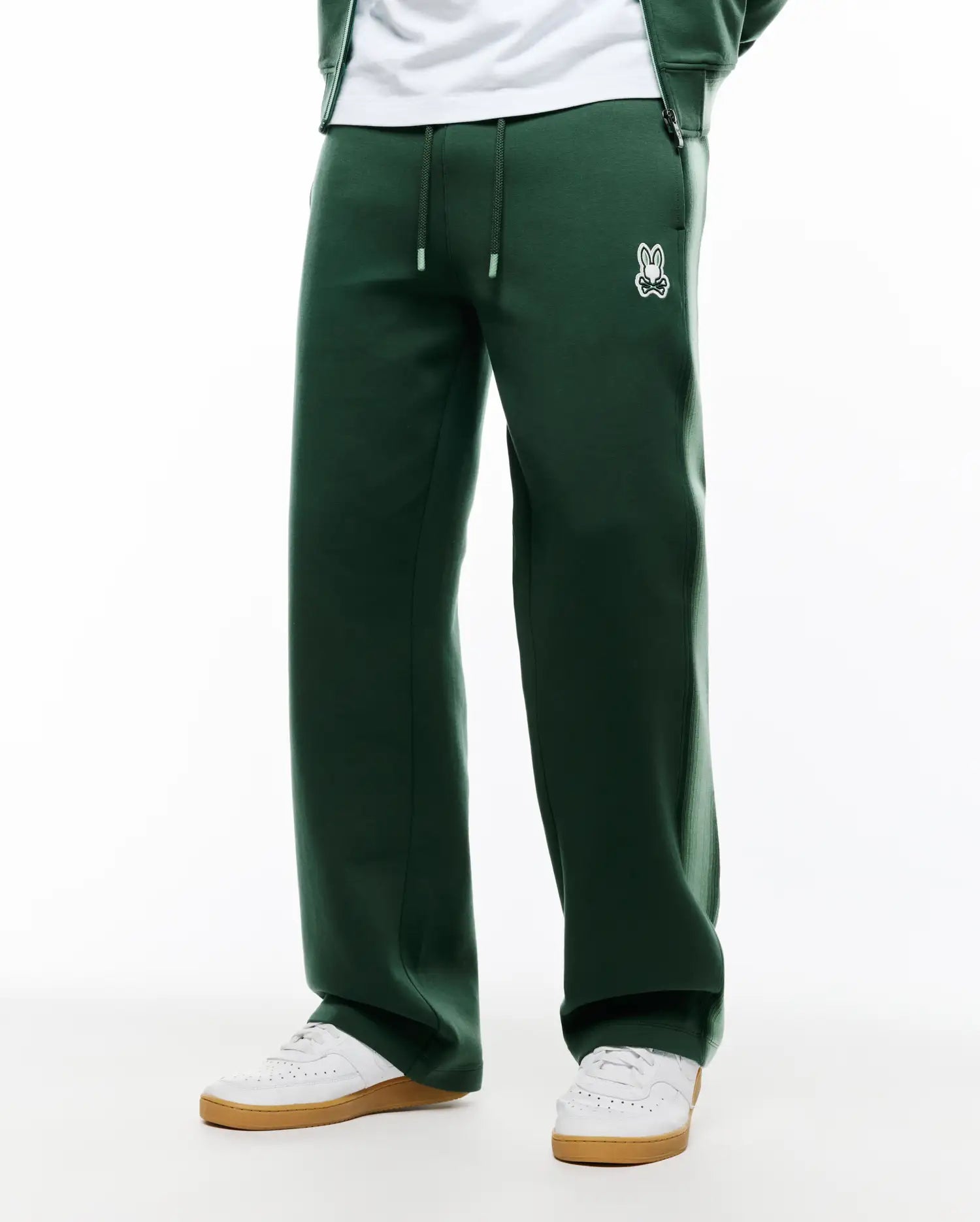 Mens Jaydon Track Pant in dark green, featuring a relaxed wide leg fit, smooth knit fabric, drawstring waist with white tips, and a small embroidered white Psycho Bunny skull logo on the left hip. (3)