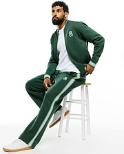Dark green Mens Jaydon Track Pant, relaxed fit, smooth knit, with light green side stripes and a small embroidered white Psycho Bunny skull logo on the left thigh, paired with a matching bomber jacket. (1)