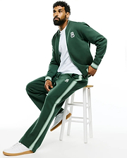 Dark green Mens Jaydon Track Pant, relaxed fit, smooth knit, with light green side stripes and a small embroidered white Psycho Bunny skull logo on the left thigh, paired with a matching bomber jacket. (1)