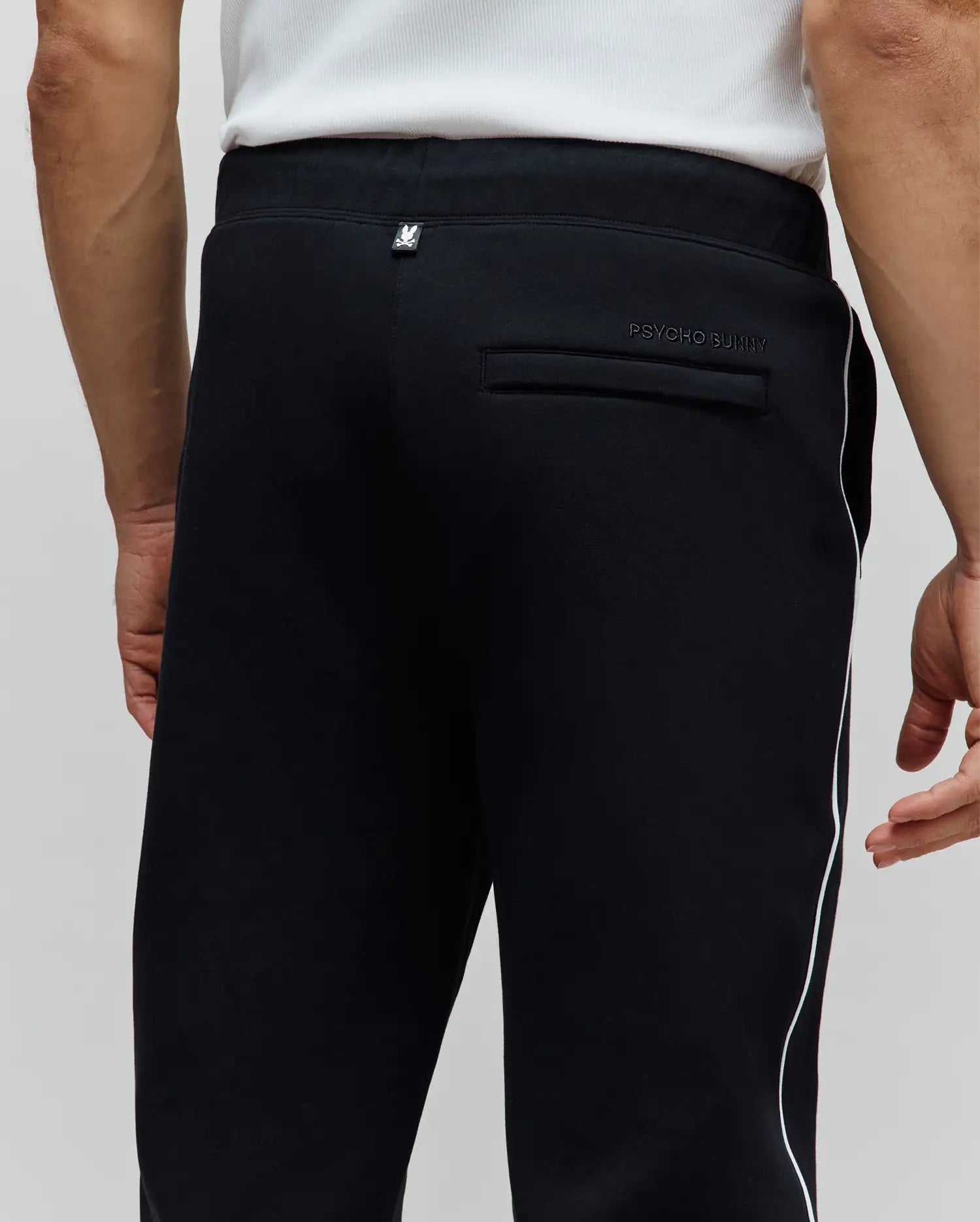 Mens Toby Track Pants, black, classic fit, smooth knit, with white Psycho Bunny skull logo on back waistband, tonal Psycho Bunny text on back pocket, and white side stripe.