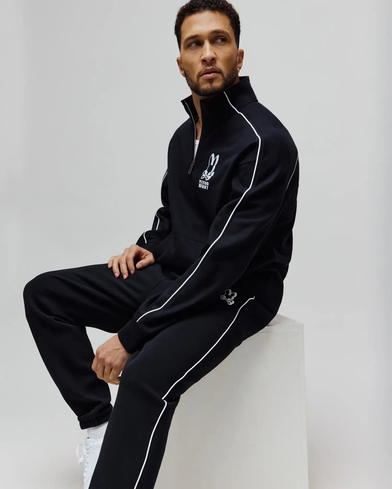 Black Psycho Bunny Mens Toby Track Pants and matching track jacket, featuring white piping details, a large embroidered bunny skull logo on the jacket's left chest, and a smaller logo on the pants' left thigh. Relaxed fit, smooth knit.