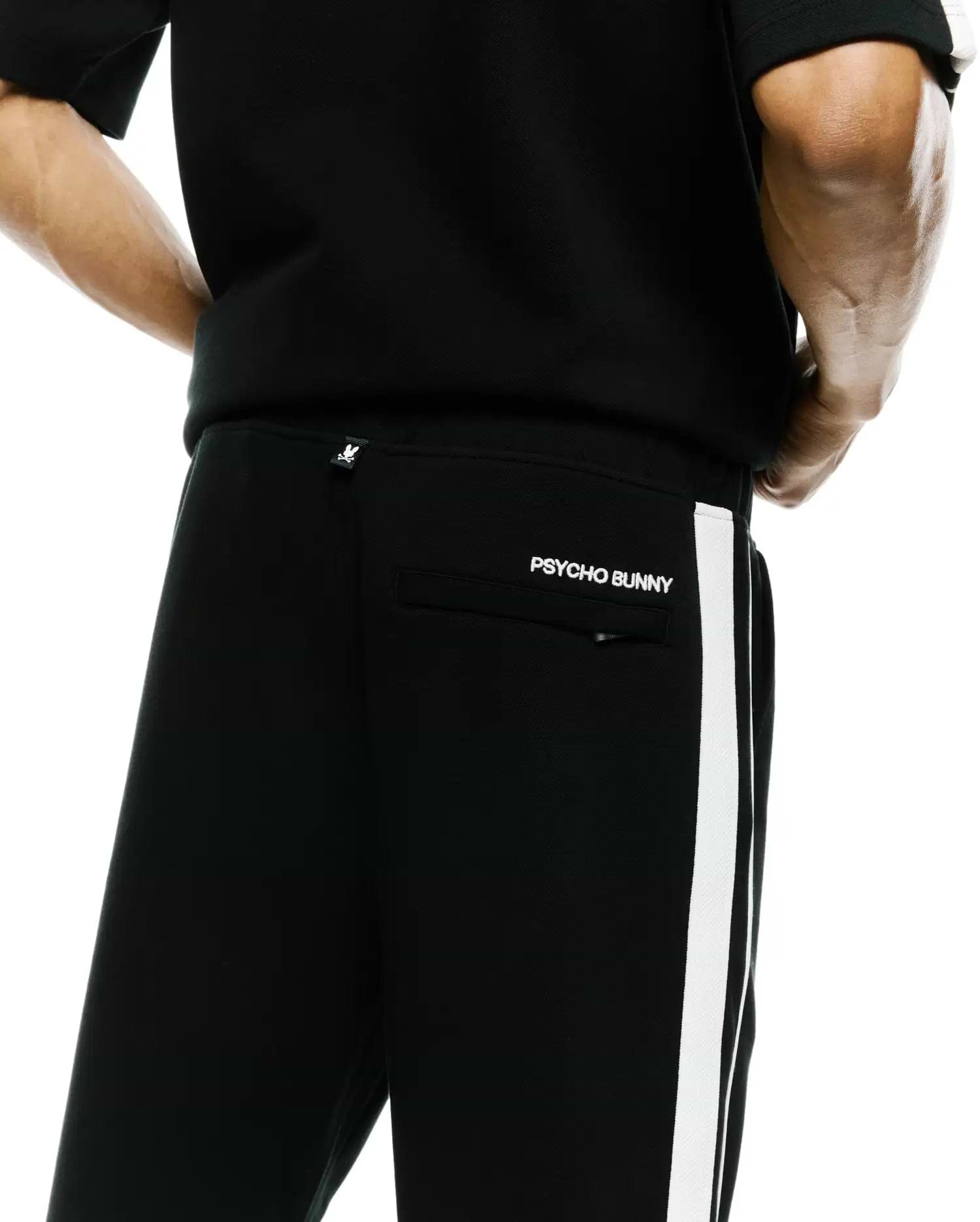 Mens Ceano Track Pant, black smooth knit with a white side stripe, featuring a white Psycho Bunny skull tag on the back waist and embroidered PSYCHO BUNNY text above the back pocket.