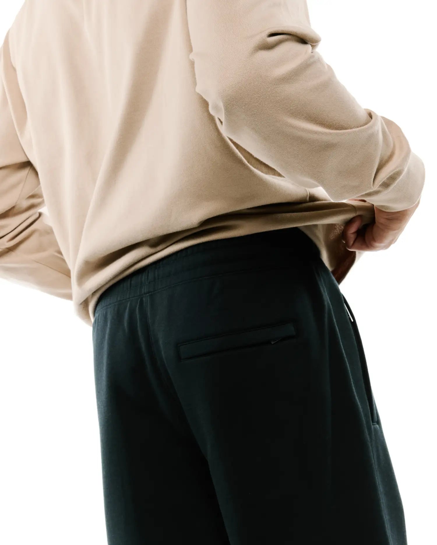 Dark charcoal Mens Terrence Brushed Fleece Sweatpant featuring a relaxed fit, elastic waistband, side pockets, and a subtle zippered back pocket, paired with a beige long-sleeve top.