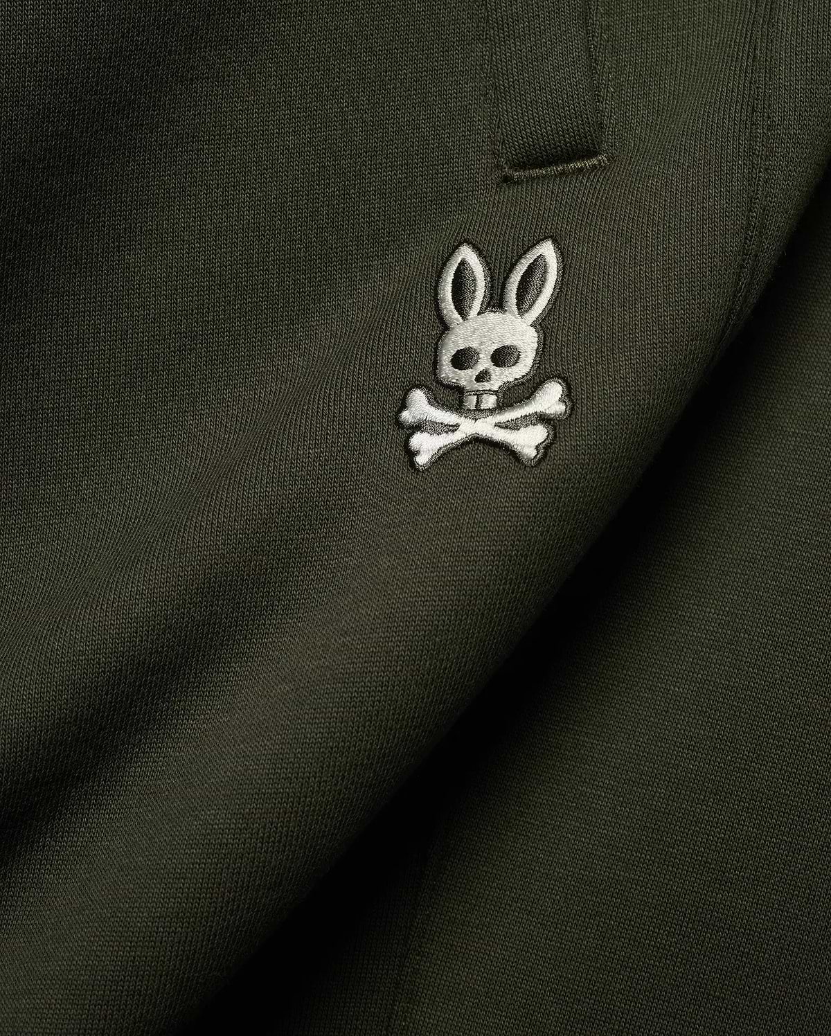 Mens Terrence Brushed Fleece Sweatpant in dark olive green, featuring a small, intricately embroidered white and silver Psycho Bunny skull logo on the upper leg, showcasing the soft brushed fleece knit texture.