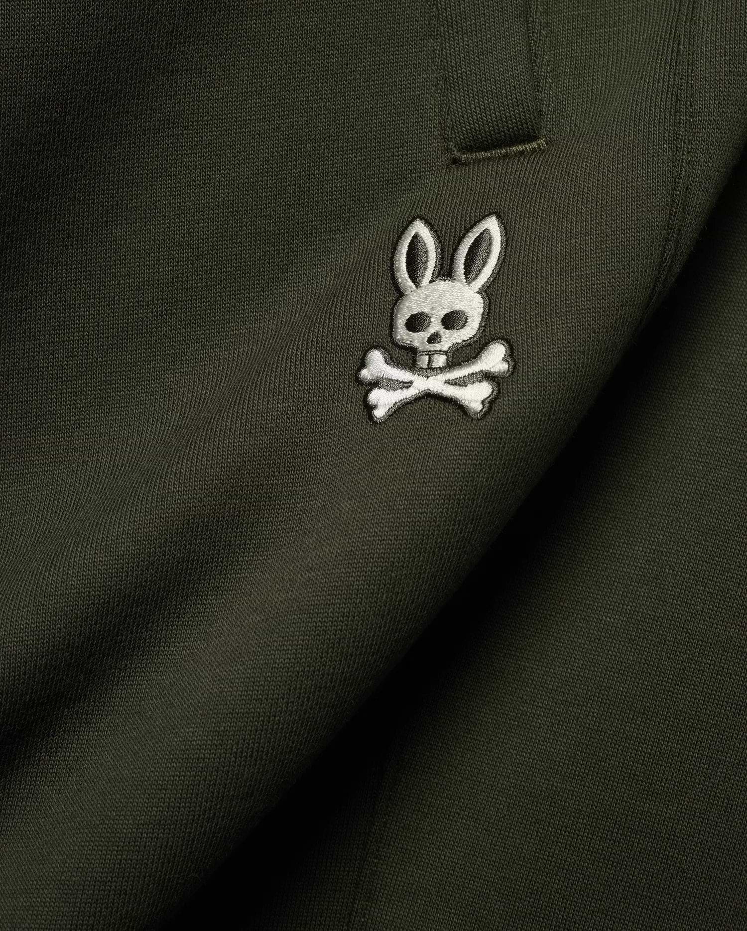 Mens Terrence Brushed Fleece Sweatpant in dark olive green, featuring a small, intricately embroidered white and silver Psycho Bunny skull logo on the upper leg, showcasing the soft brushed fleece knit texture.