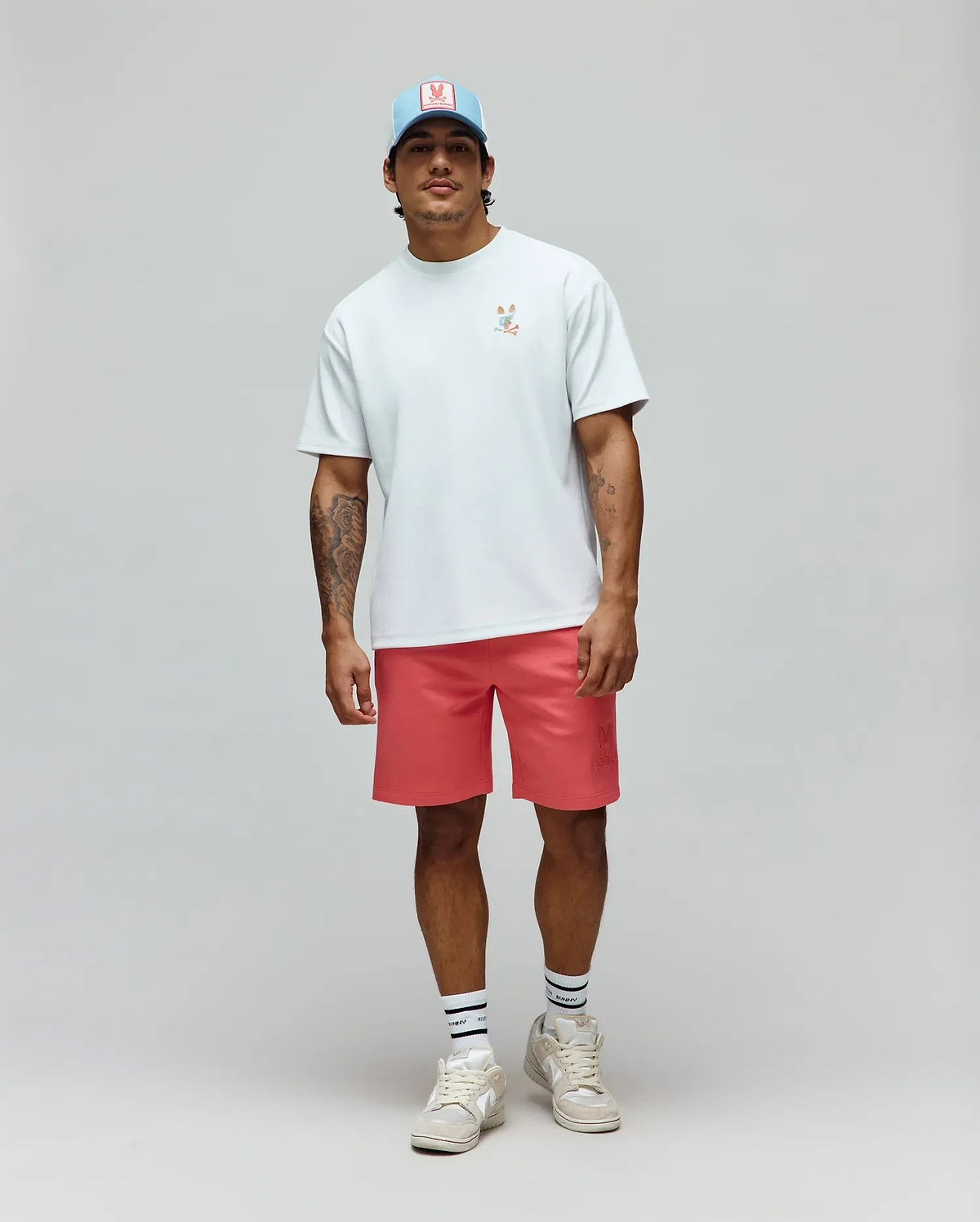 Pale blue jersey t-shirt with a small, colorful embroidered Psycho Bunny skull logo on the chest, paired with coral Mens Barry Short featuring an embossed bunny skull on the left thigh, and a light blue trucker hat with a pink Psycho Bunny patch. (4)