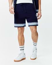 Mens Giles Knit Short 7, a navy textured knit short with a drawstring waist, featuring light blue and white horizontal stripes at the hem and a small embroidered white Psycho Bunny skull logo on the left thigh.
