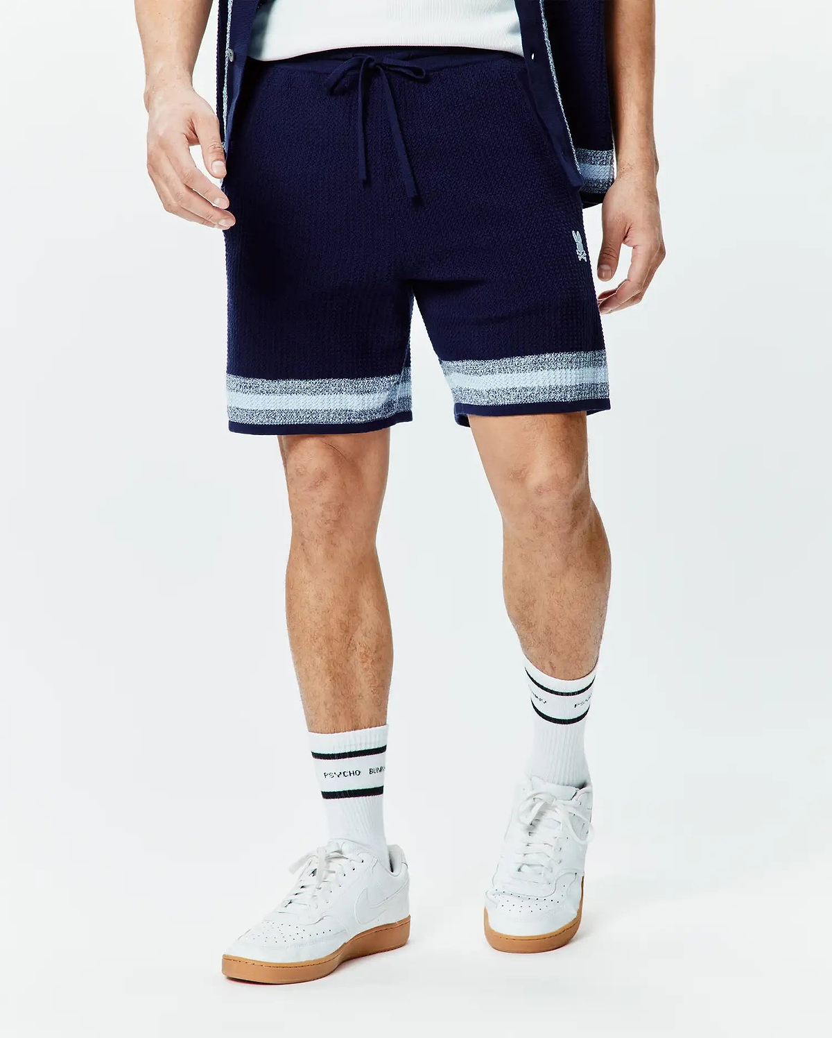 Mens Giles Knit Short 7, a navy textured knit short with a drawstring waist, featuring light blue and white horizontal stripes at the hem and a small embroidered white Psycho Bunny skull logo on the left thigh.