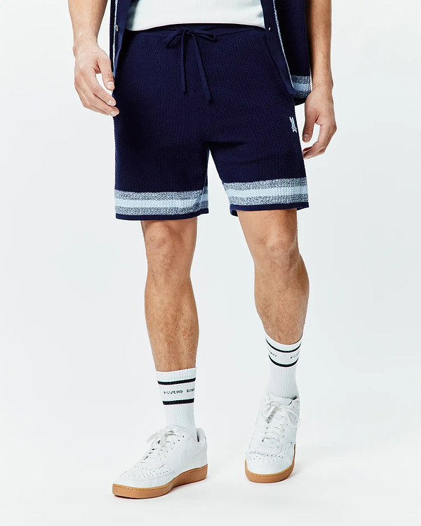 Mens Giles Knit Short 7, a navy textured knit short with a drawstring waist, featuring light blue and white horizontal stripes at the hem and a small embroidered white Psycho Bunny skull logo on the left thigh.