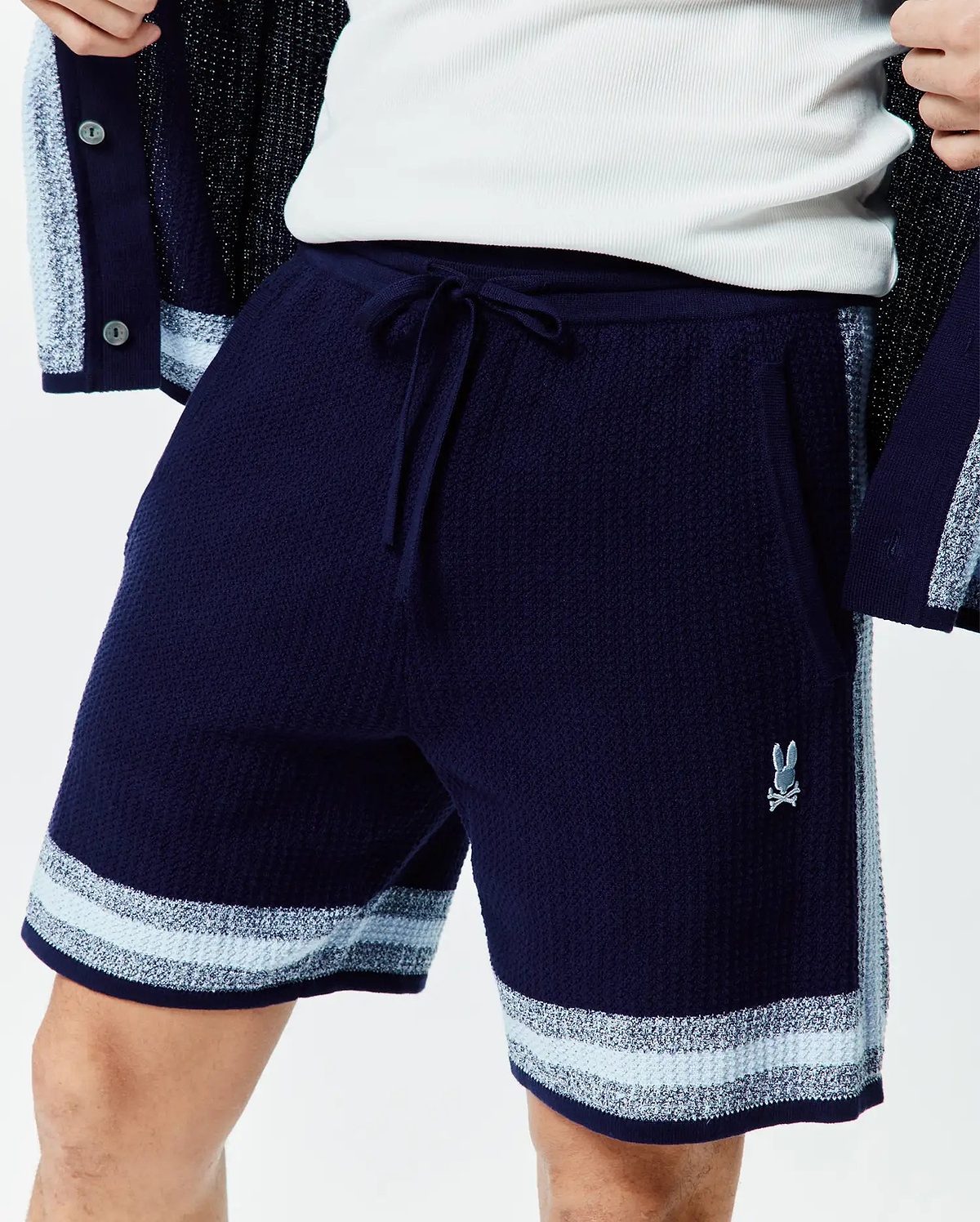 Mens Giles Knit Short 7. Navy blue textured knit shorts with a drawstring waist, light blue and white striped hem, side panel, and small embroidered Psycho Bunny logo on the right leg.