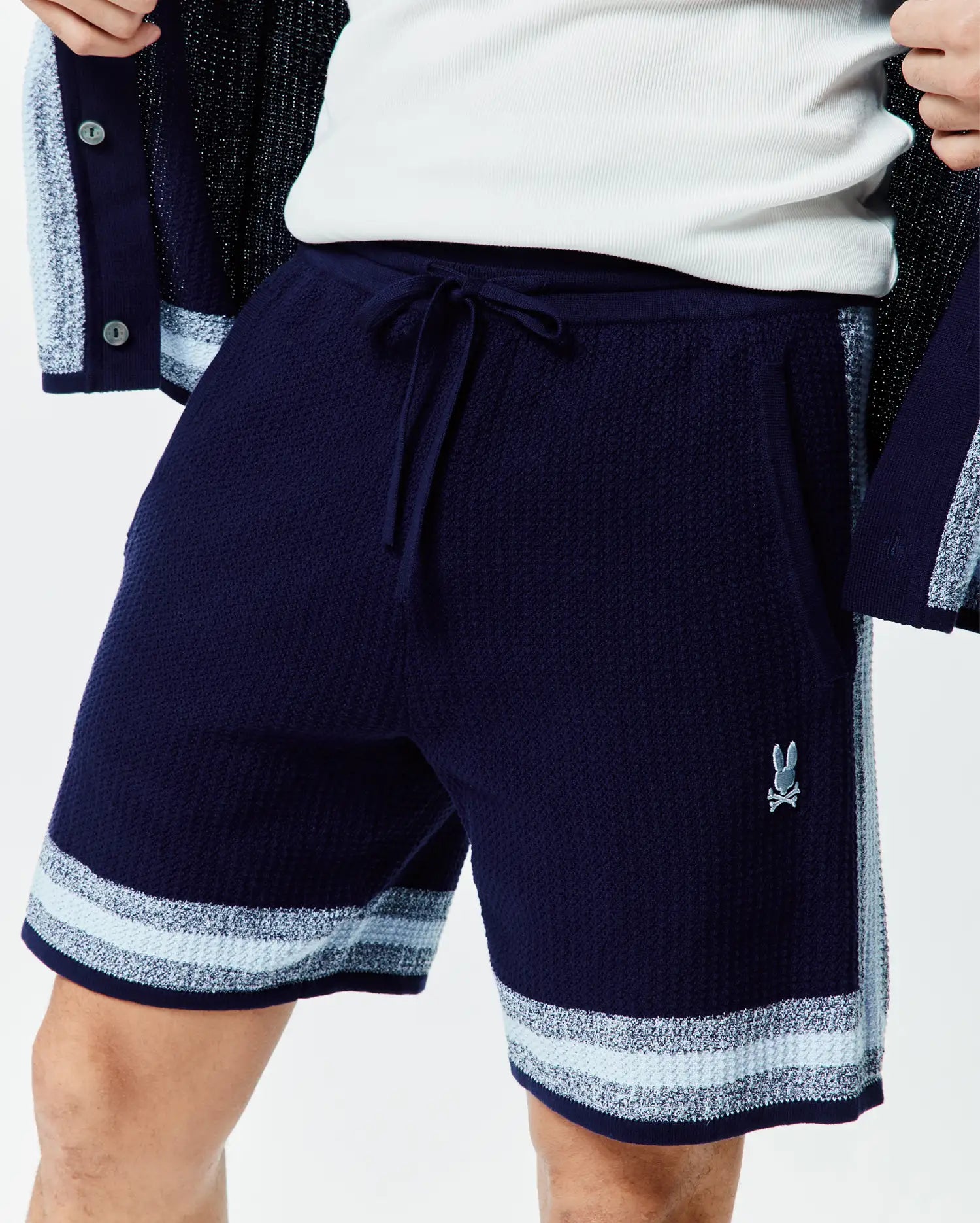 Mens Giles Knit Short 7. Navy blue textured knit shorts with a drawstring waist, light blue and white striped hem, side panel, and small embroidered Psycho Bunny logo on the right leg.