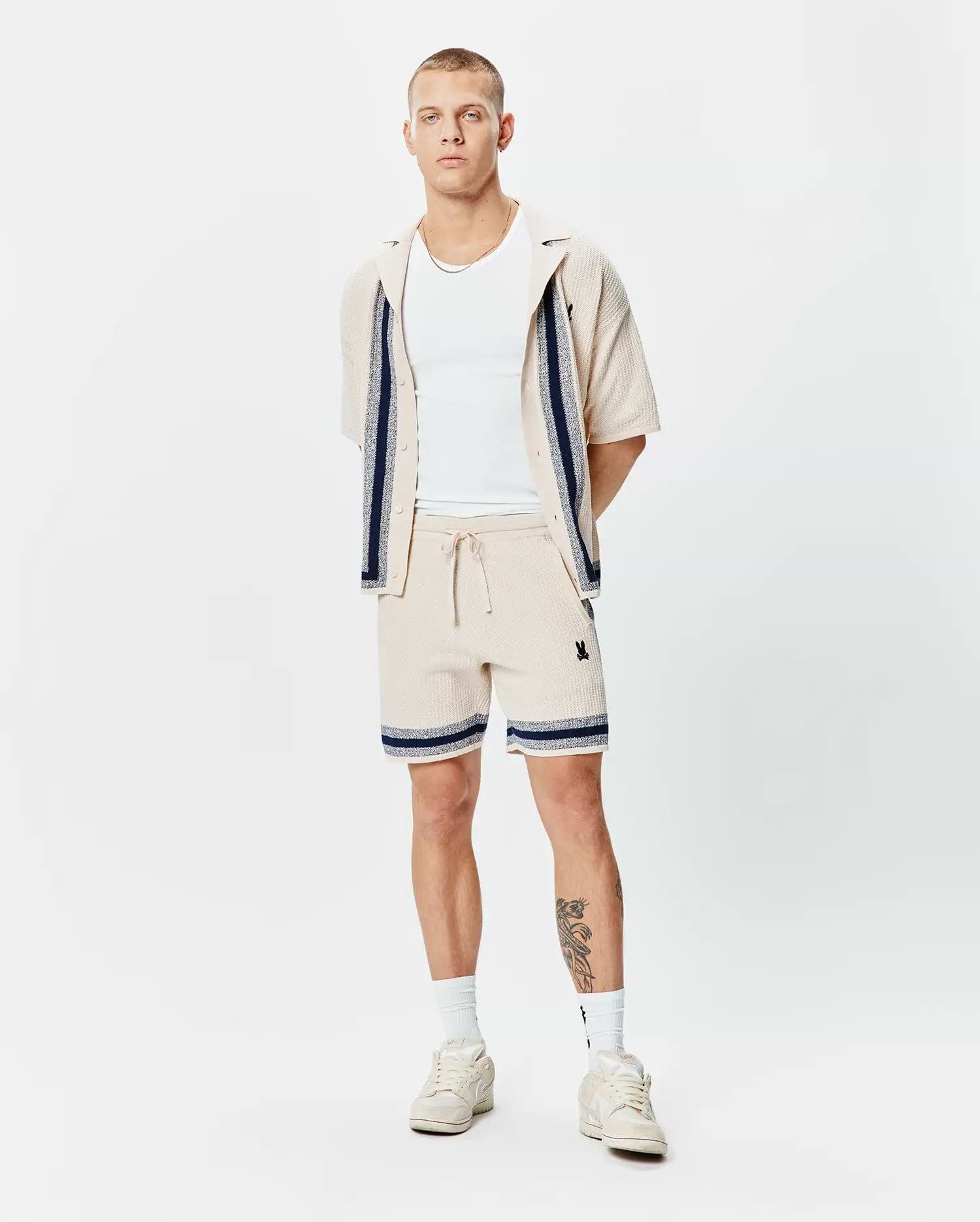 Mens Giles Knit Short 7 and matching collared shirt in beige textured knit with navy and white striped accents and small embroidered Psycho Bunny skull logo on shorts' thigh.
