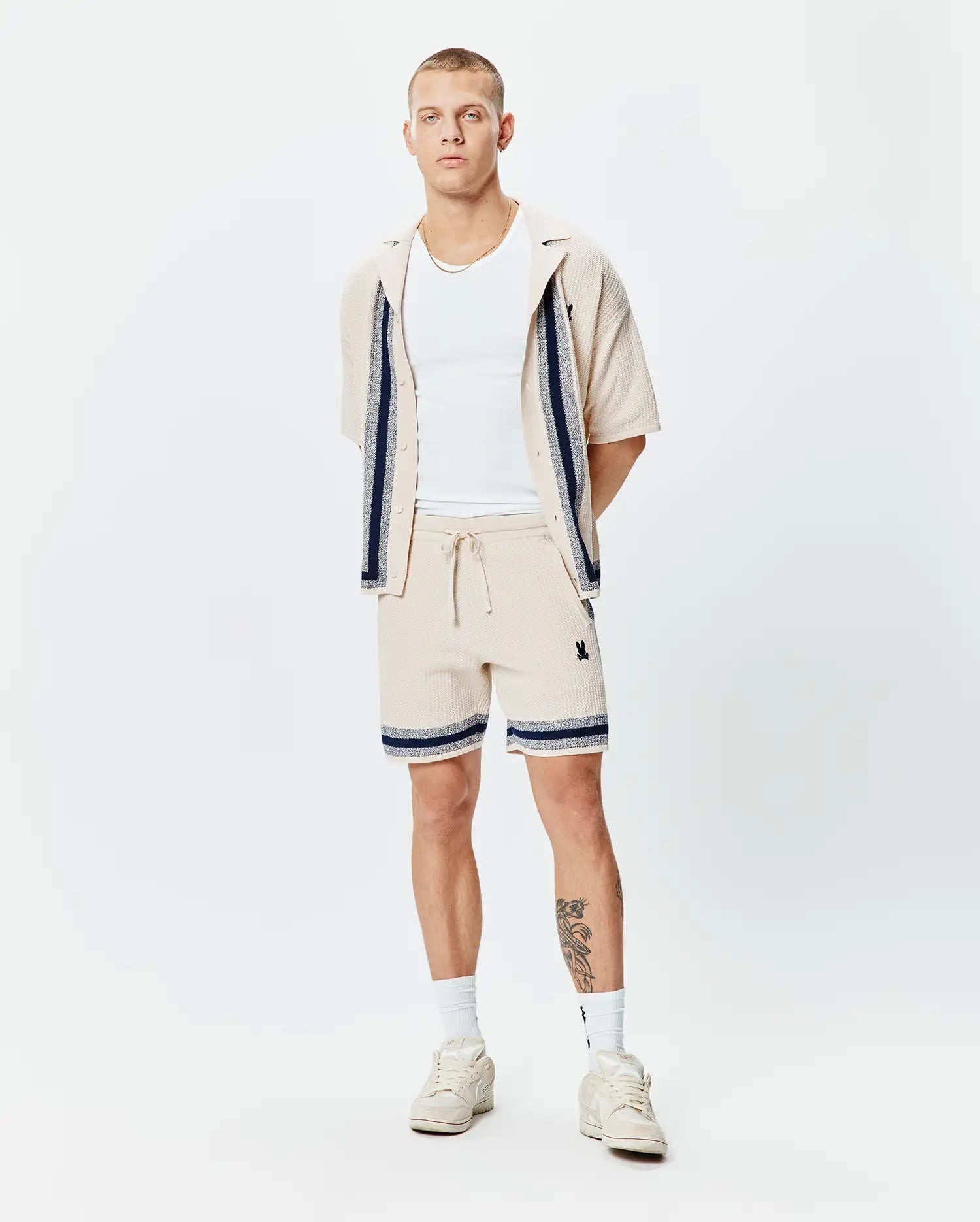 Mens Giles Knit Short 7 and matching collared shirt in beige textured knit with navy and white striped accents and small embroidered Psycho Bunny skull logo on shorts' thigh.