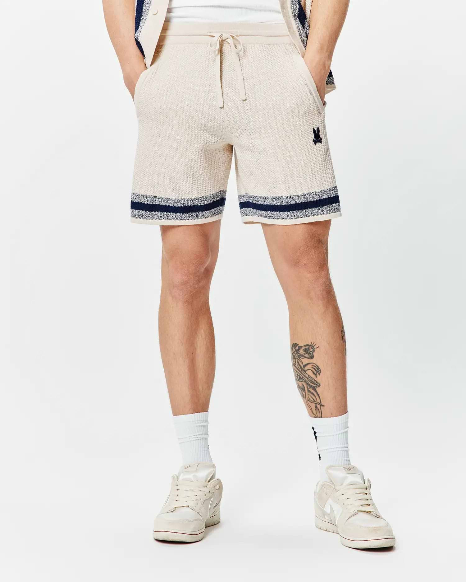 Mens Giles Knit Short 7, a cream textured knit short with navy and white striped hem, featuring a small embroidered Psycho Bunny skull logo on the left thigh and a drawstring waist.