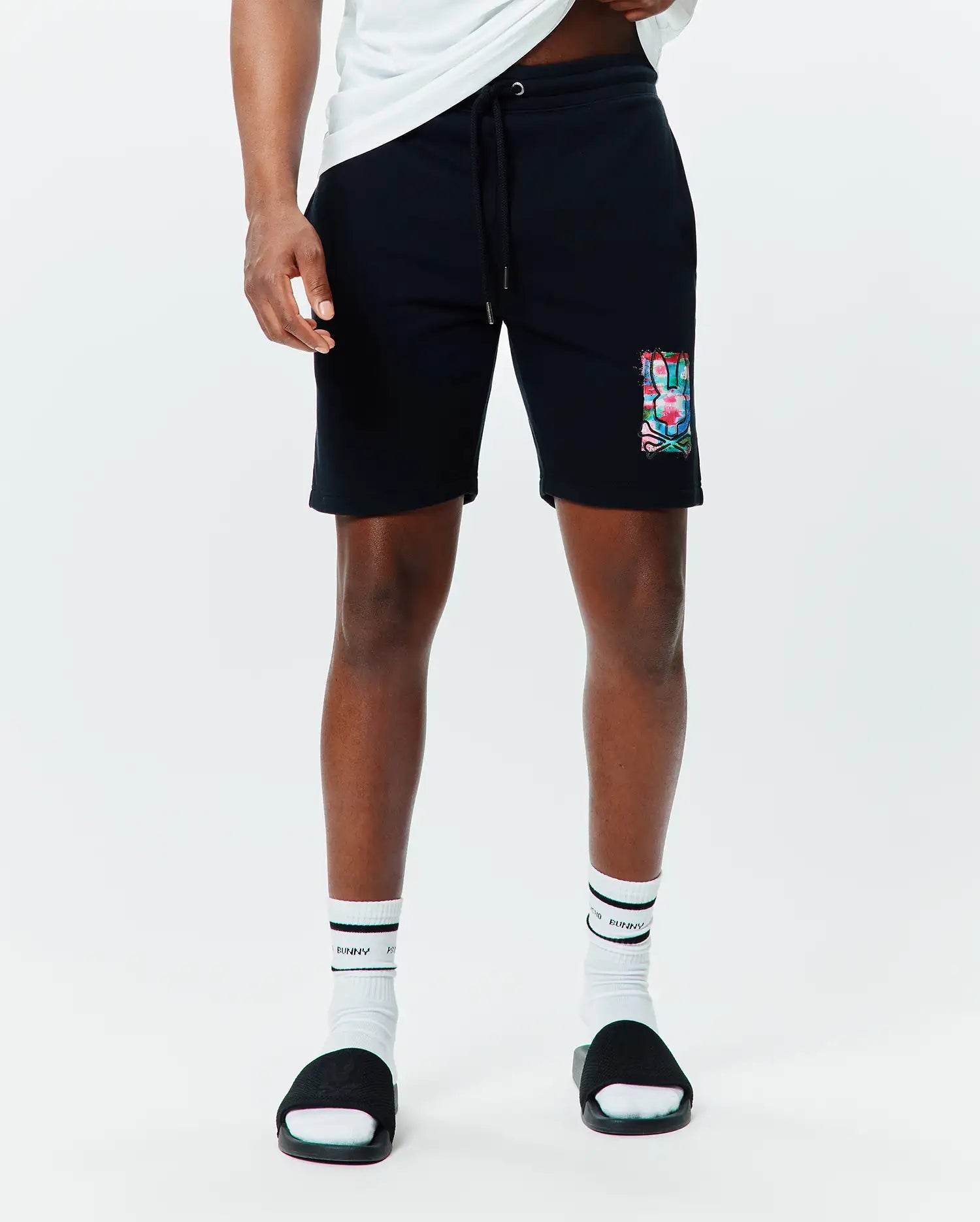 Mens Isaac Print Short, black relaxed fit French terry shorts with a drawstring waist, featuring a large, vibrant multi-color Psycho Bunny skull graphic print on the left thigh.