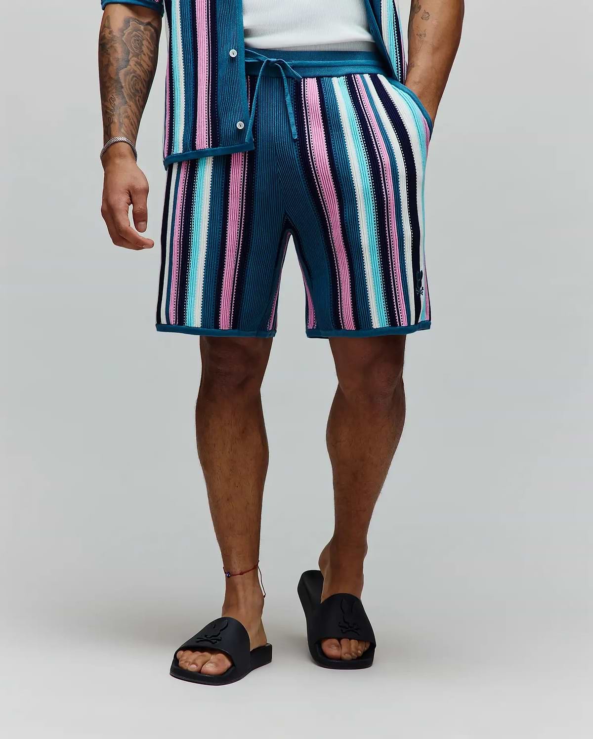 Mens Cliff Knit Short, relaxed fit, vibrant multi-color vertical stripes in teal, pink, light blue, white, and navy ribbed knit, with a small embroidered Psycho Bunny skull logo on the lower left leg.