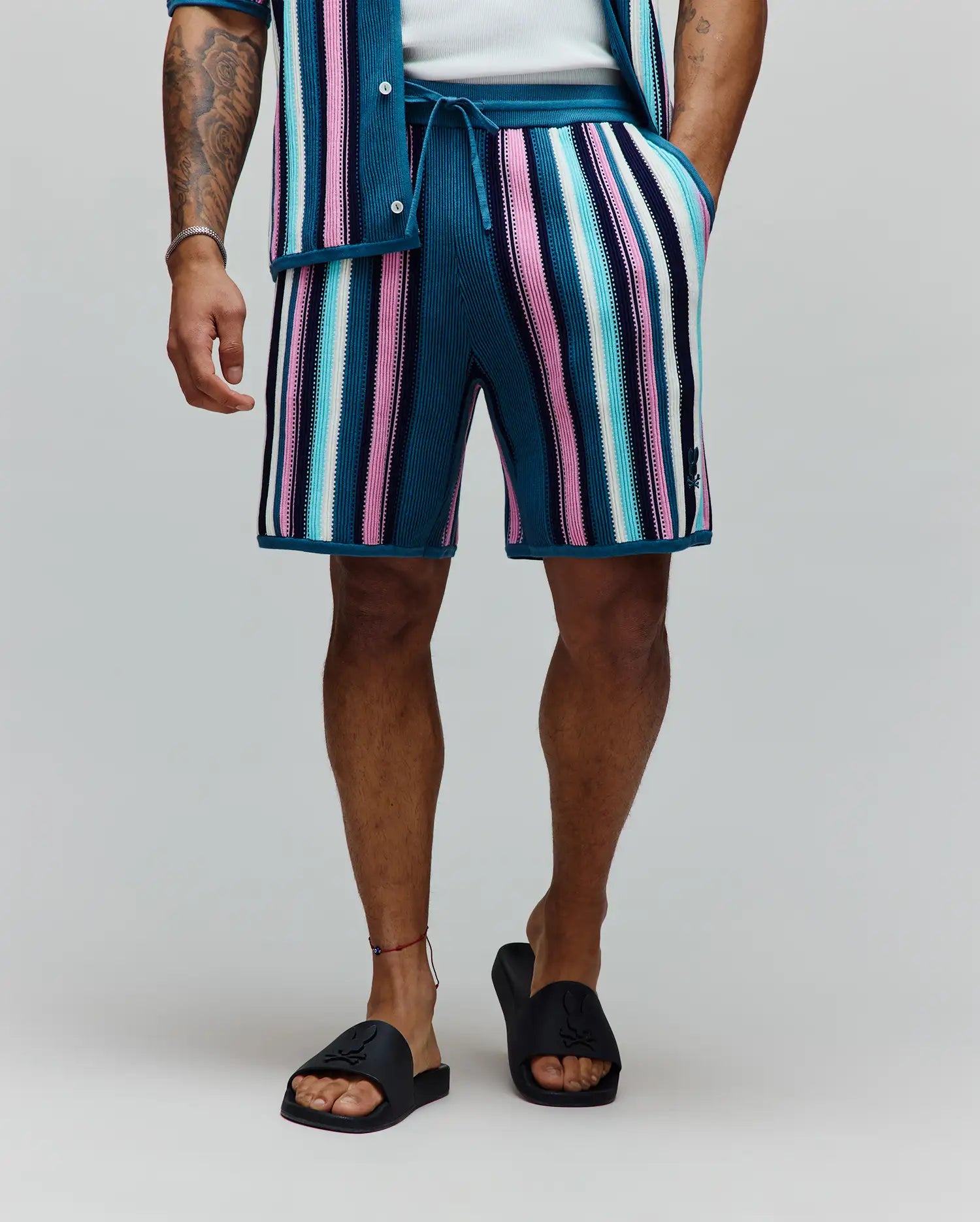 Mens Cliff Knit Short, relaxed fit, vibrant multi-color vertical stripes in teal, pink, light blue, white, and navy ribbed knit, with a small embroidered Psycho Bunny skull logo on the lower left leg.