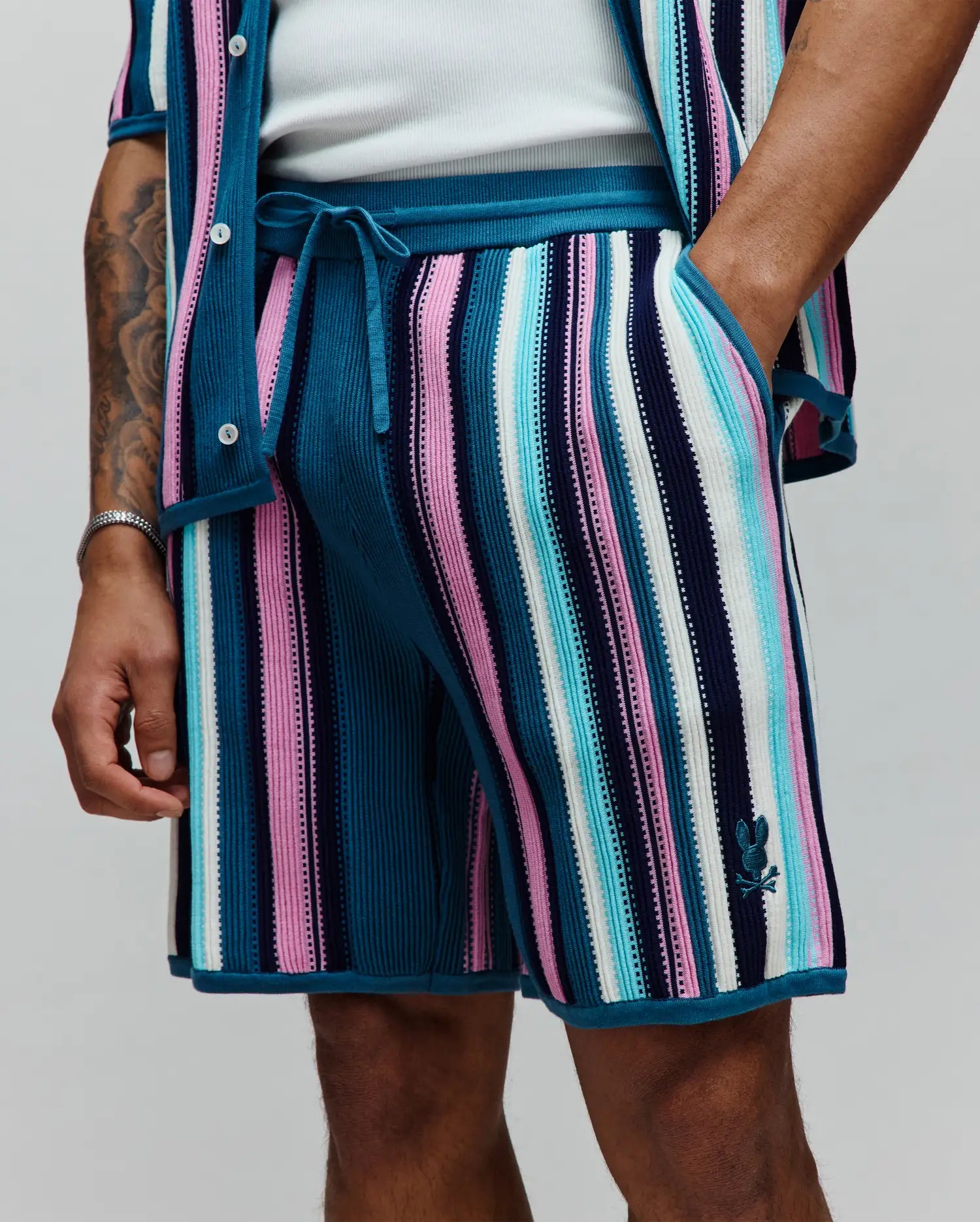 Mens Cliff Knit Short with multi-color vertical stripes in teal, pink, white, light blue, and navy blue, featuring a ribbed knit texture, drawstring waist, and a small embroidered Psycho Bunny skull logo on the lower right leg.