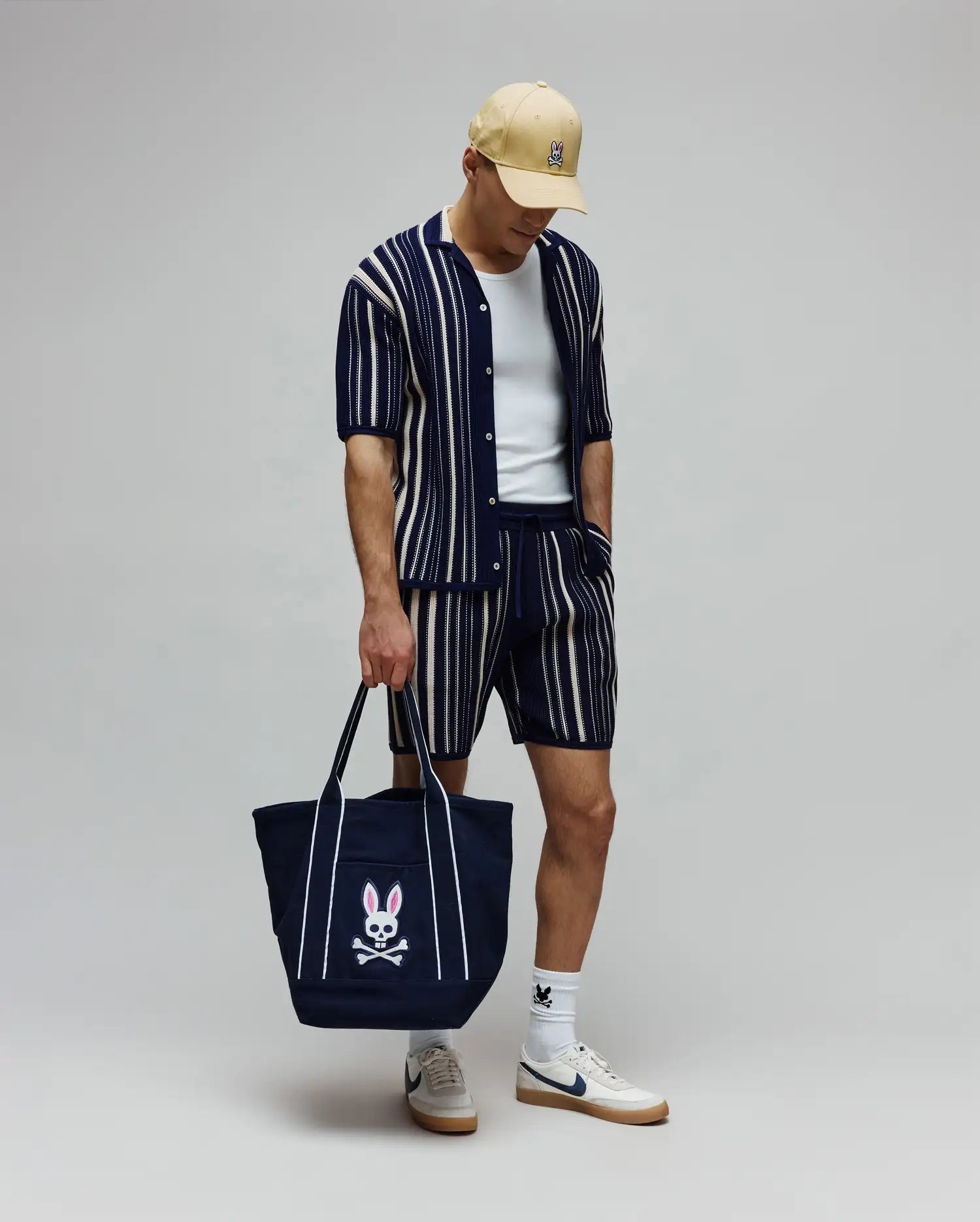 Relaxed fit navy and cream striped knit short-sleeve shirt and matching Mens Cliff Knit Short, complemented by a tan cap and white socks, all featuring the iconic Psycho Bunny skull logo.