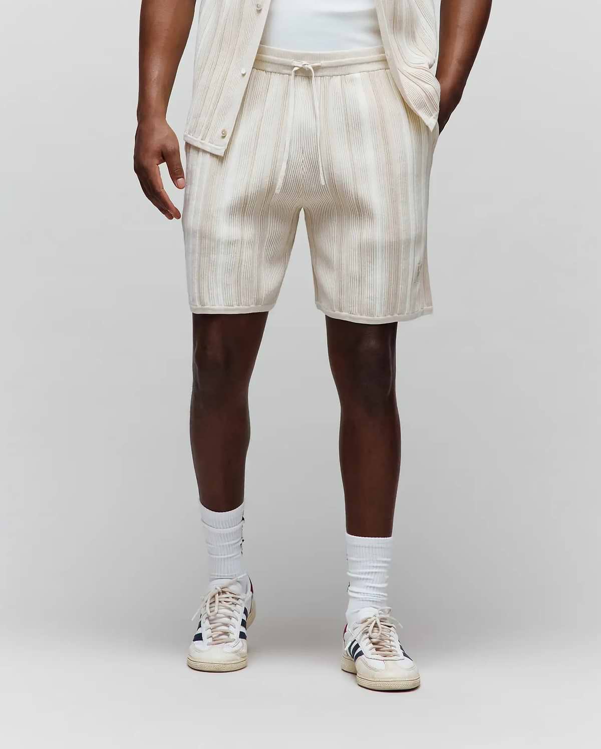 Mens Cliff Knit Short in cream with subtle beige vertical stripes, featuring a relaxed fit, ribbed knit texture, drawstring waist, and a small embroidered Psycho Bunny skull logo on the lower left leg.