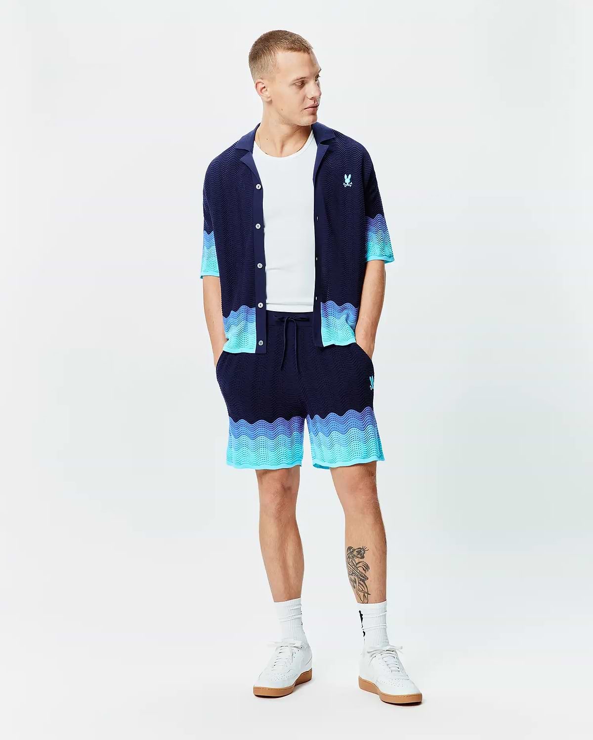Navy blue knit short-sleeve shirt and Mens Holden Knit Short 6, featuring a wavy knit texture with light blue to turquoise gradient accents, and small embroidered Psycho Bunny logos on the chest and right thigh.