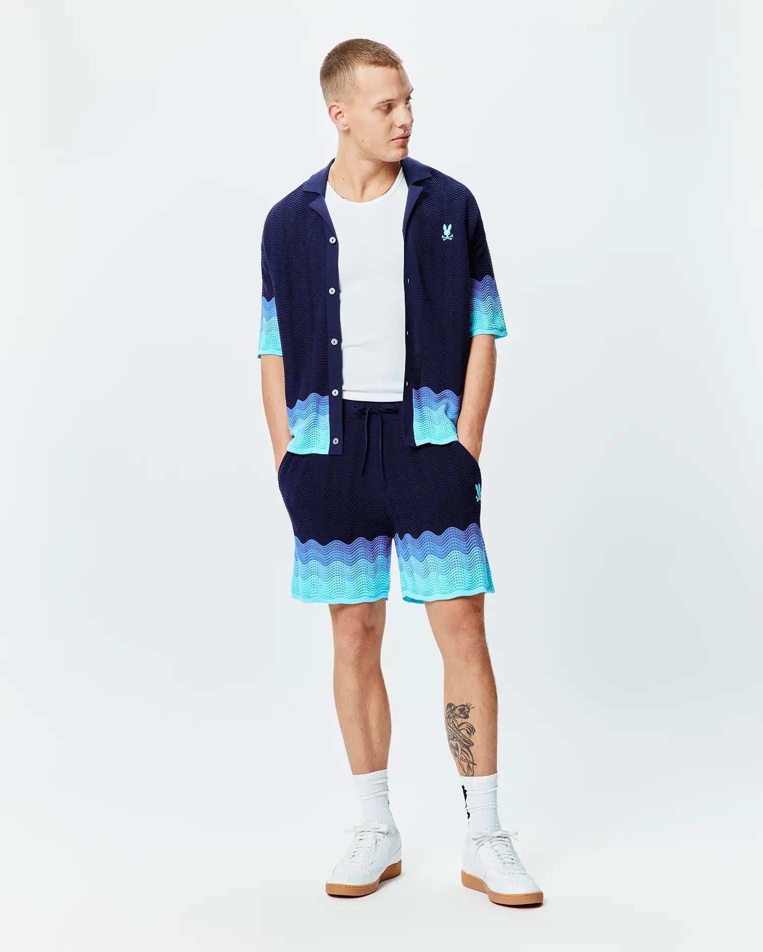 Navy blue knit short-sleeve shirt and Mens Holden Knit Short 6, featuring a wavy knit texture with light blue to turquoise gradient accents, and small embroidered Psycho Bunny logos on the chest and right thigh.
