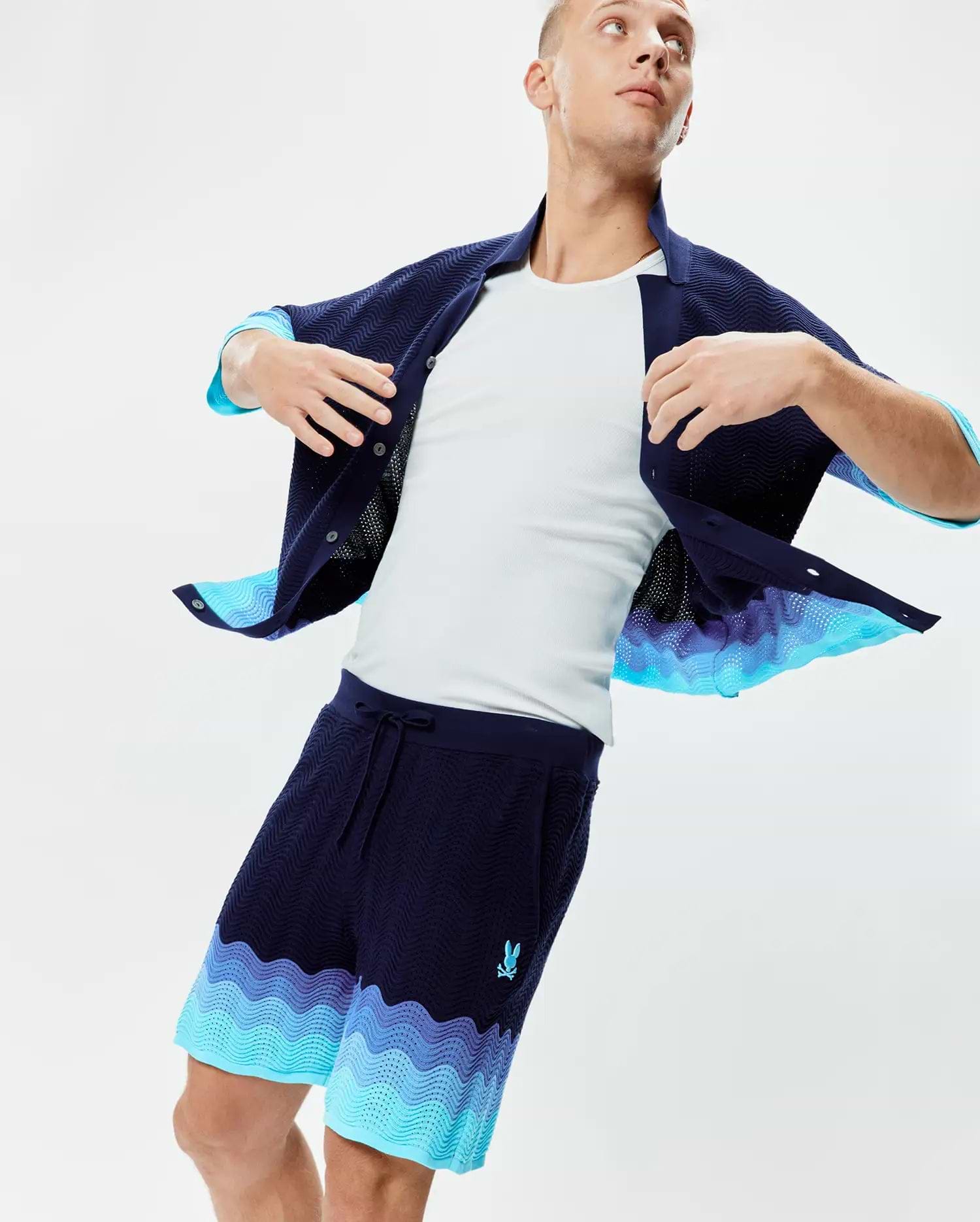 Mens Holden Knit Short 6 in navy blue with a wavy knit texture, featuring a vibrant blue and purple gradient wave pattern at the hem and a small embroidered Psycho Bunny logo on the left thigh.