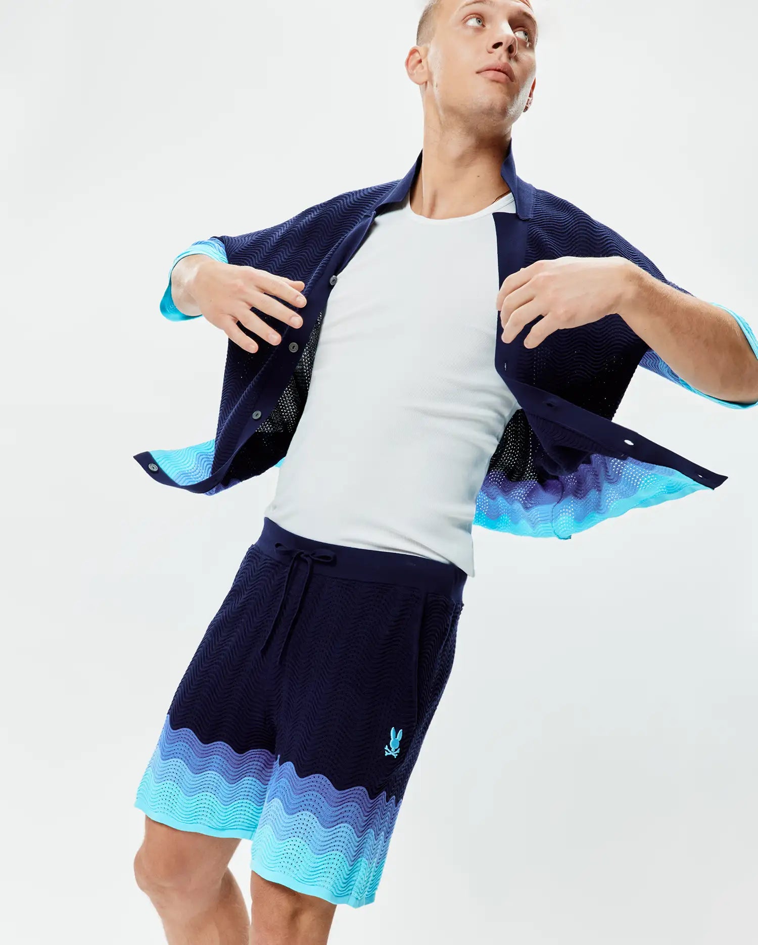 Mens Holden Knit Short 6 in navy blue with a wavy knit texture, featuring a vibrant blue and purple gradient wave pattern at the hem and a small embroidered Psycho Bunny logo on the left thigh.