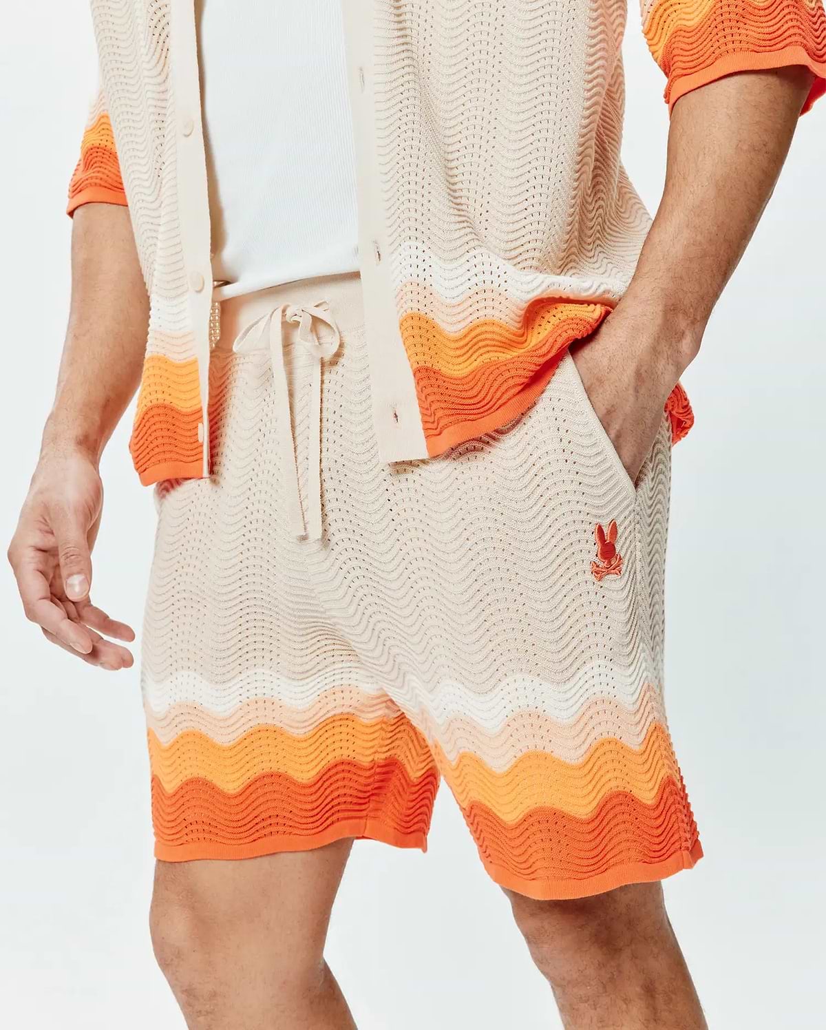 Mens Holden Knit Short 6, a relaxed fit cream open knit short with a wavy texture, featuring a drawstring waist, bold orange and white wavy stripe accents, and a small embroidered orange Psycho Bunny skull logo on the left thigh.