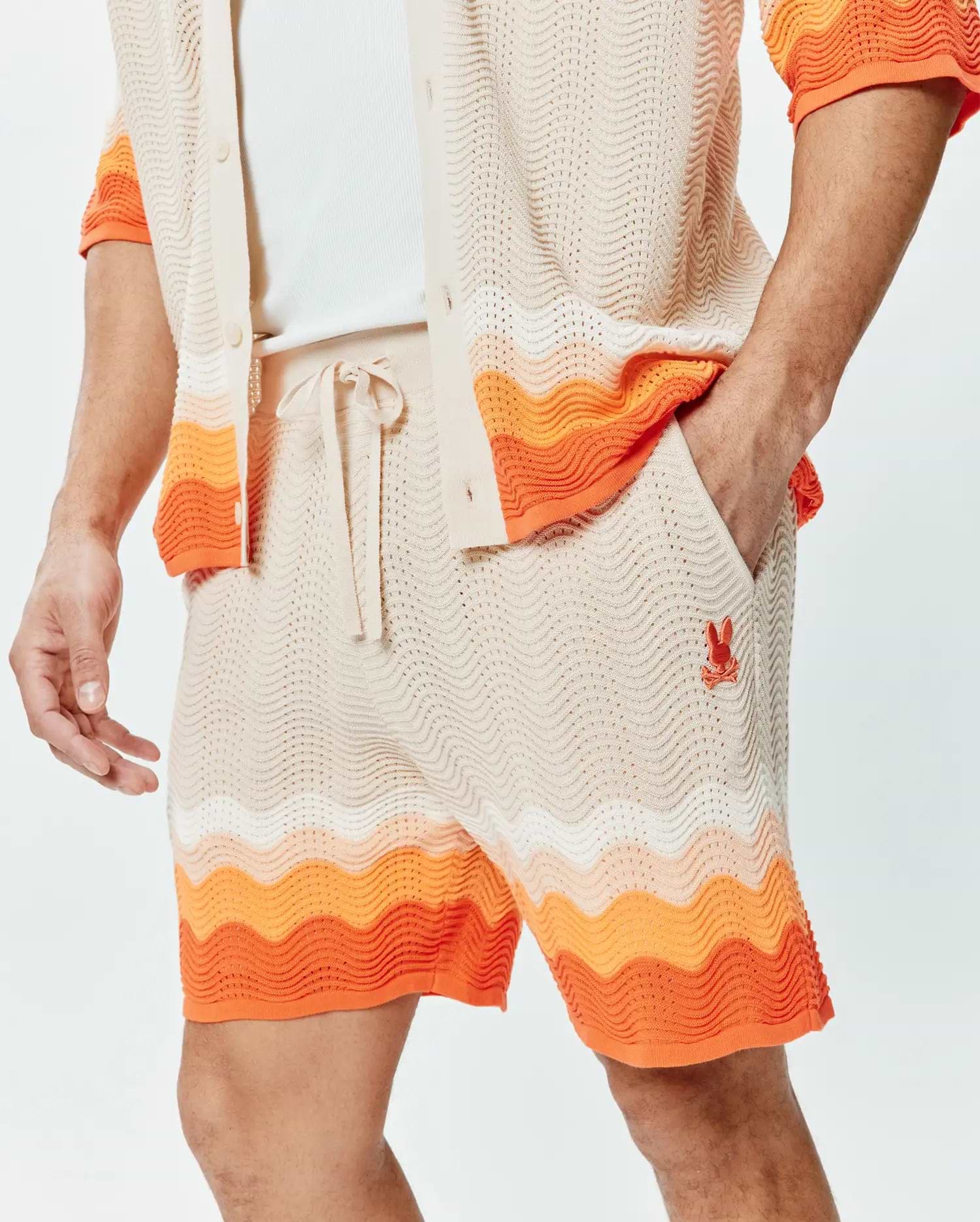 Mens Holden Knit Short 6, a relaxed fit cream open knit short with a wavy texture, featuring a drawstring waist, bold orange and white wavy stripe accents, and a small embroidered orange Psycho Bunny skull logo on the left thigh.
