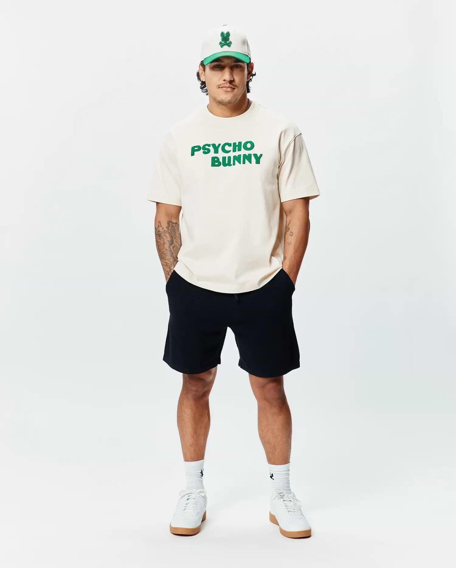 Cream classic fit jersey t-shirt with bold green PSYCHO BUNNY chest text, paired with black Mens Devon Knit Short, a cream and green Psycho Bunny skull logo cap, and white logo socks.