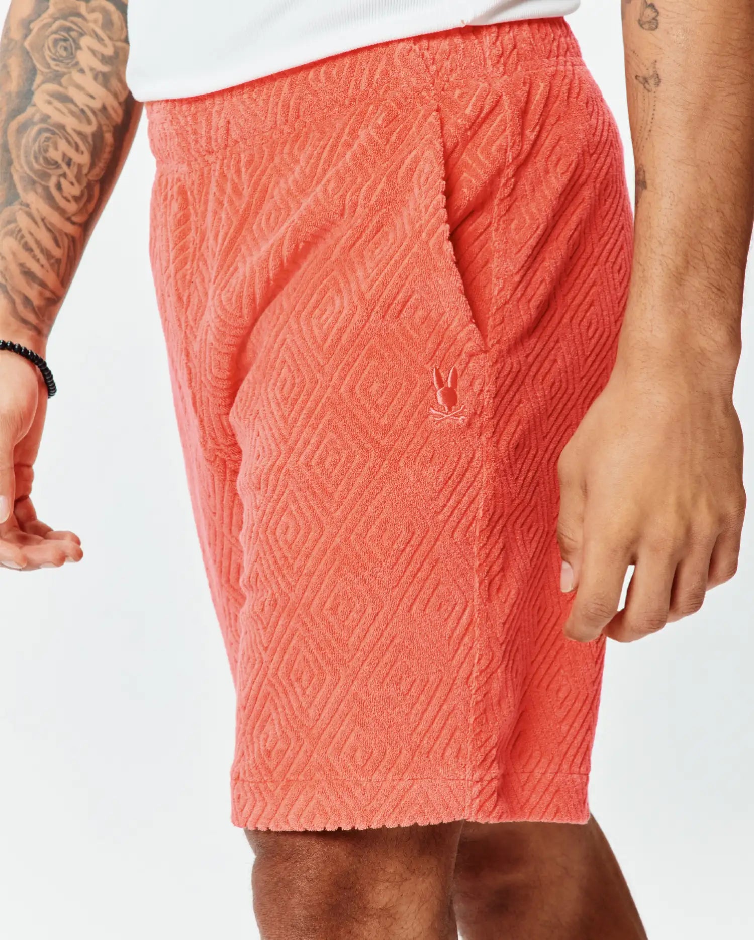 Vibrant coral terry cloth Mens Tristan Short with a relaxed fit, featuring an all-over embossed diamond pattern and a small embroidered Psycho Bunny skull logo on the left thigh.