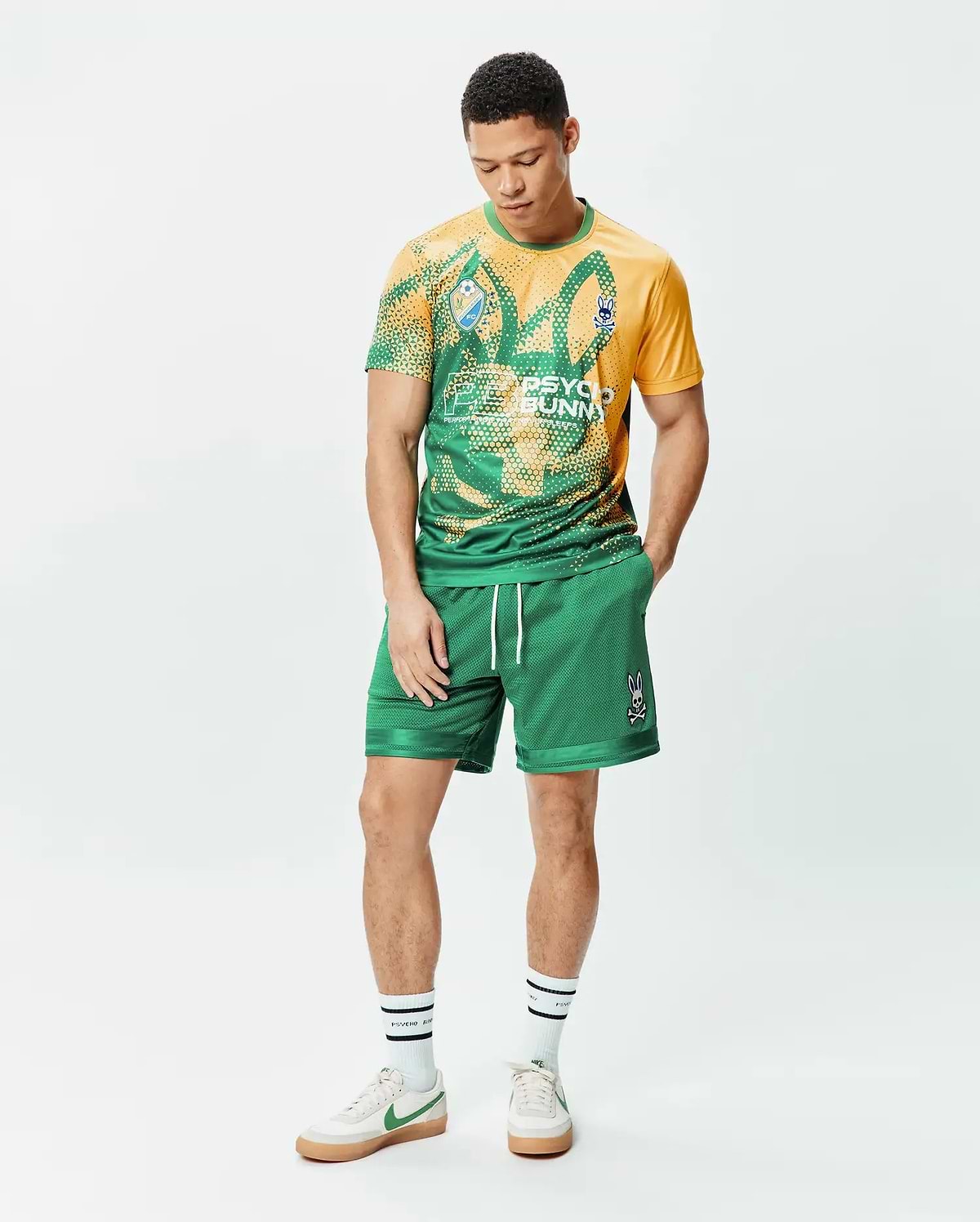 Mens Joshua Soccer Short 7 1/4 in green mesh with white drawstrings and an embroidered Psycho Bunny skull logo on the right leg, paired with a yellow and green graphic t-shirt featuring a large bunny skull print and small sleeve logo. (4)