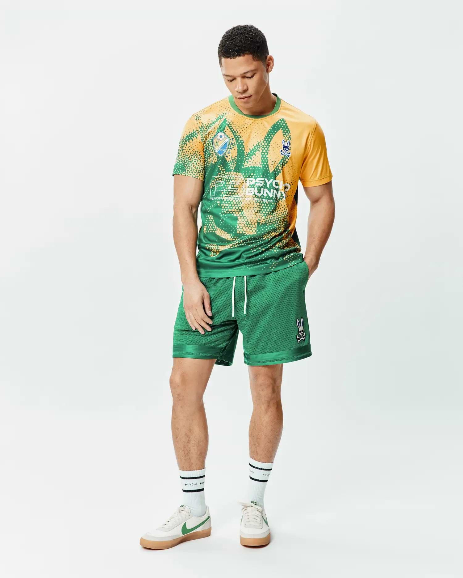 Mens Joshua Soccer Short 7 1/4 in green mesh with white drawstrings and an embroidered Psycho Bunny skull logo on the right leg, paired with a yellow and green graphic t-shirt featuring a large bunny skull print and small sleeve logo. (4)
