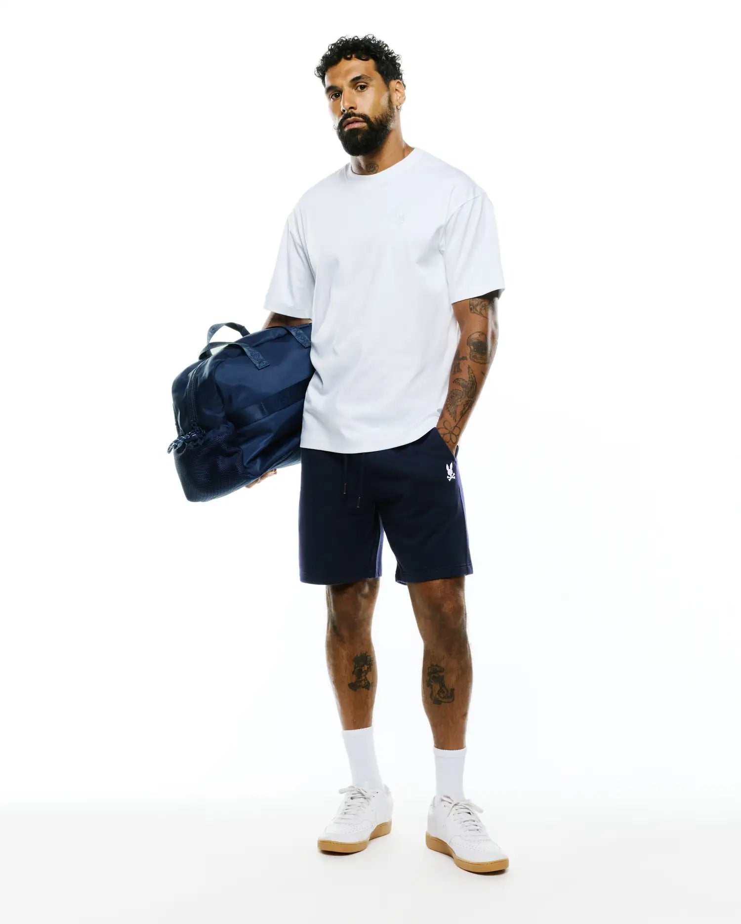 White relaxed fit t-shirt with a small embroidered Psycho Bunny logo on the chest, paired with navy Mens Gabe French Terry Sweatshort featuring a drawstring waist and a white embroidered bunny skull logo on the left leg. (5)